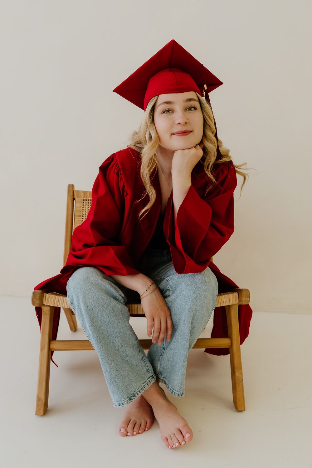 Senior girl in red graduation cap and gown sitting barefoot on wooden chair, posing thoughtfully.