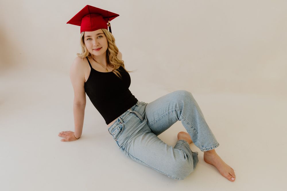 A smiling blonde graduate wearing a red cap poses seated on the floor in jeans and a black top.