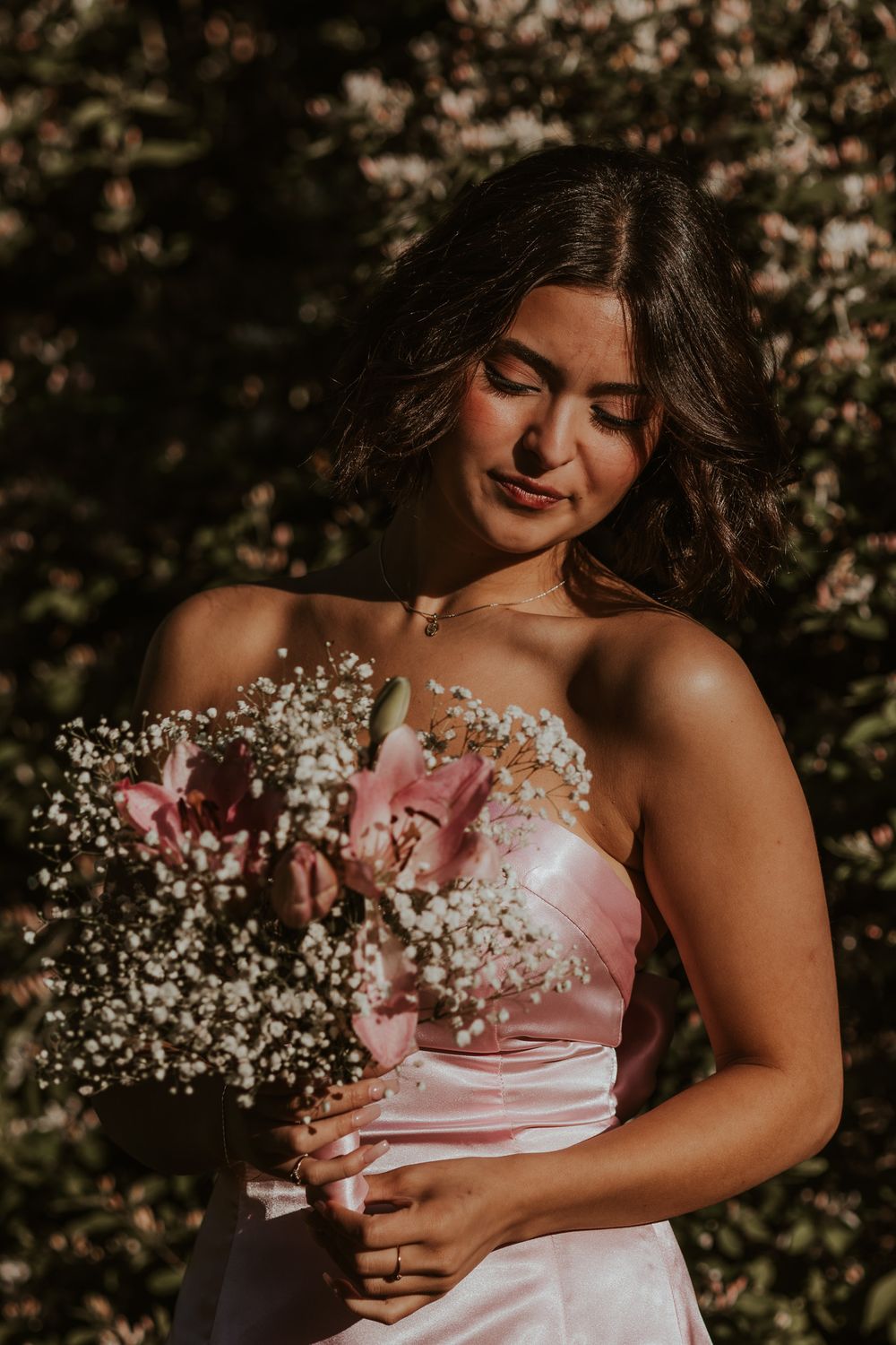 Young woman in pink strapless gown holding a bouquet of pink lilies and baby's breath outdoors.