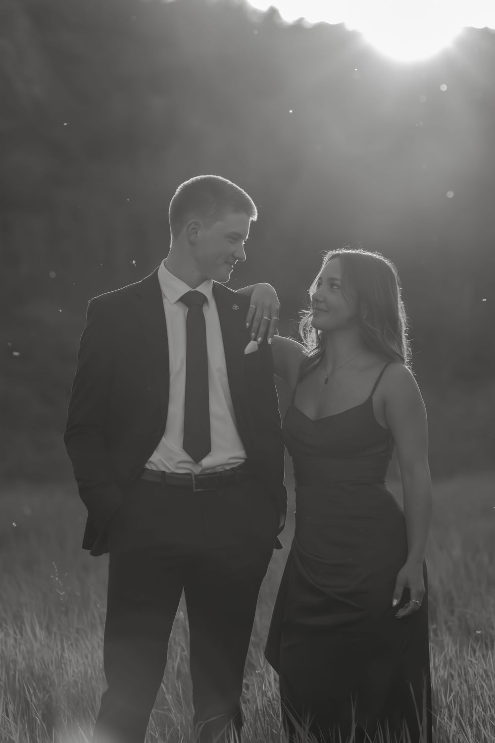 Black and white photo of a couple in formal attire smiling at each other in a sunlit field.