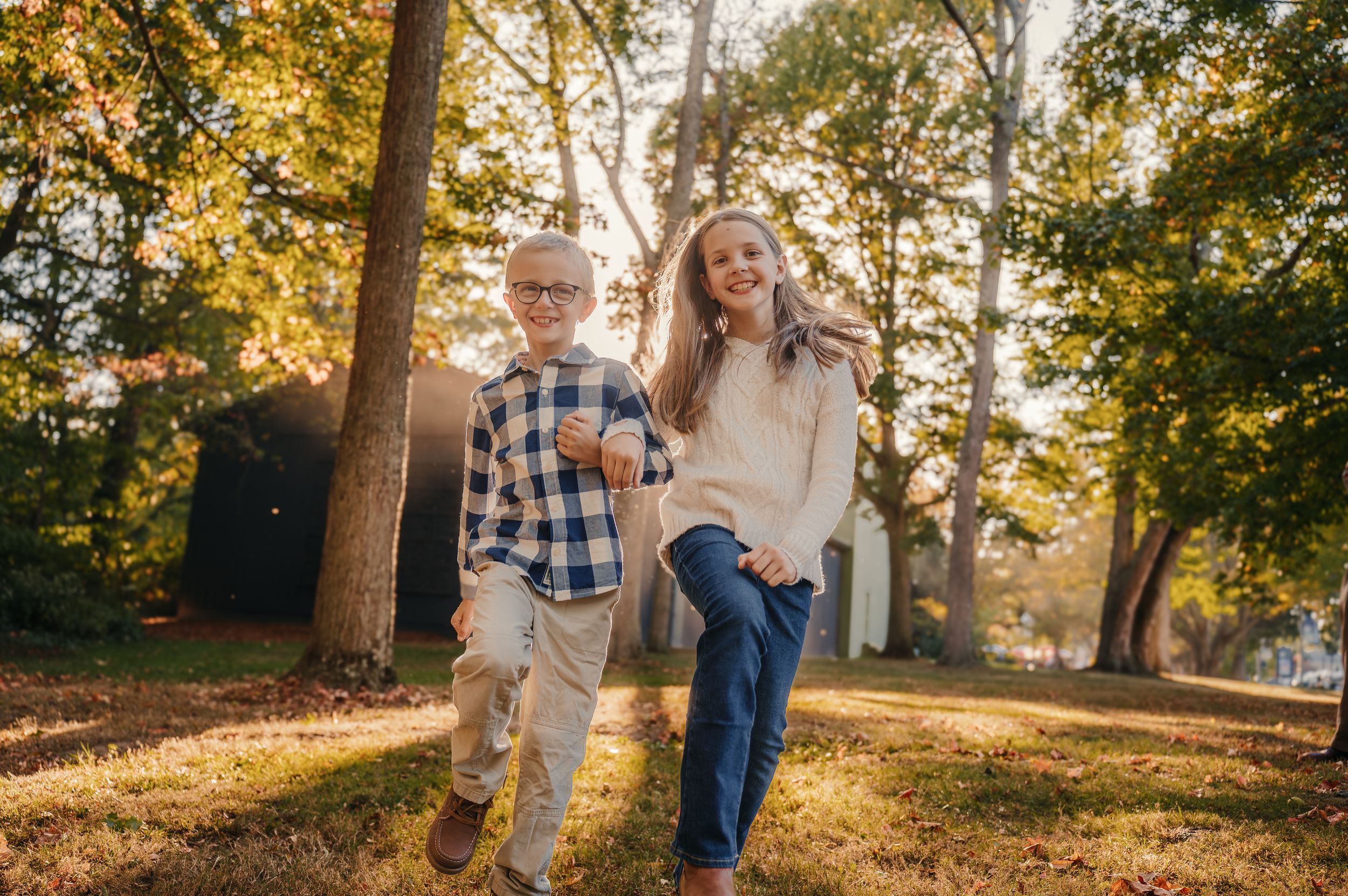 Two children running and laughing outdoors in a sunlit autumn park surrounded by golden trees.
