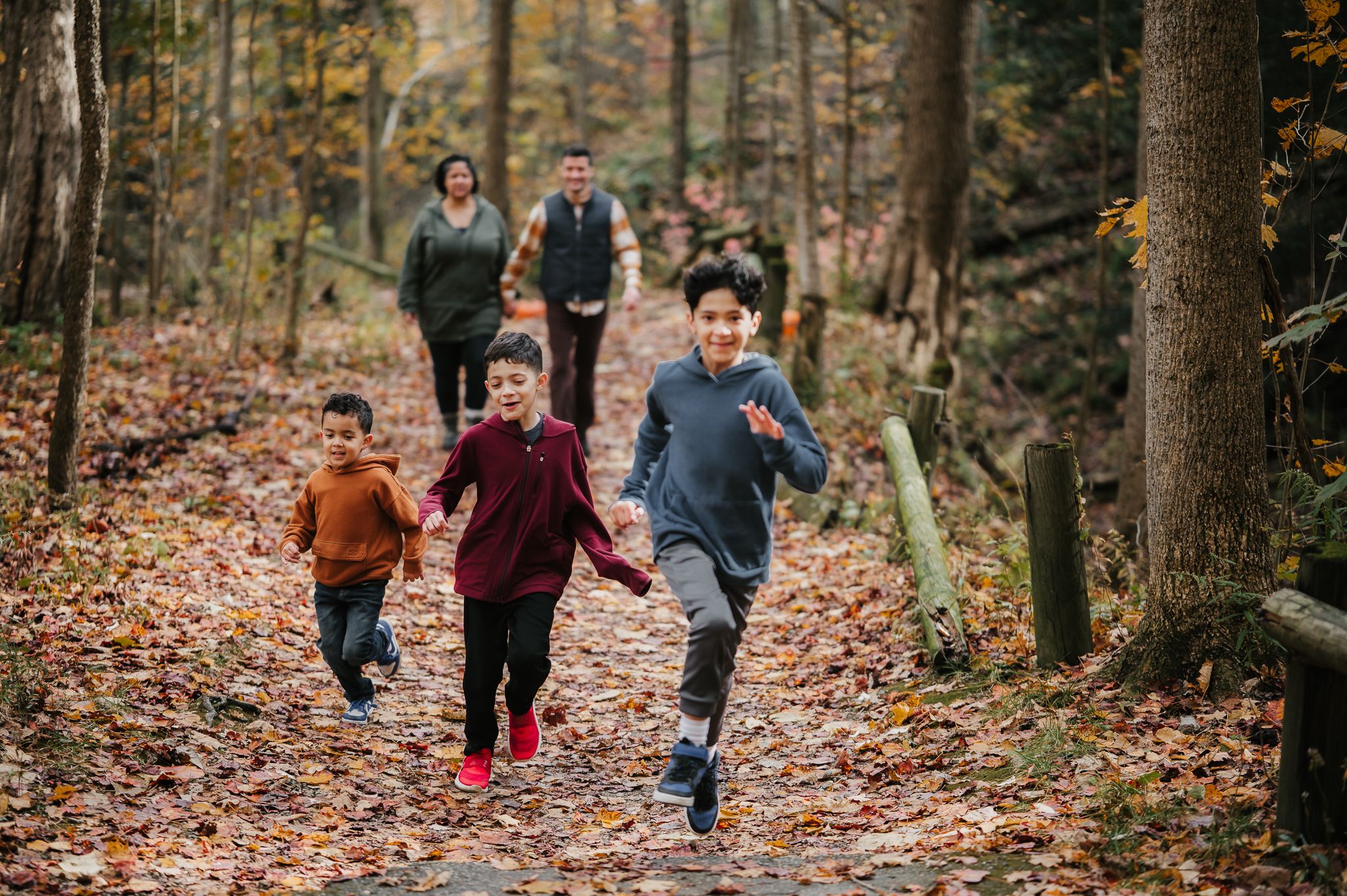 Family hiking on a leaf-covered forest trail in autumn, with children running ahead of parents among colorful fall foliage.
