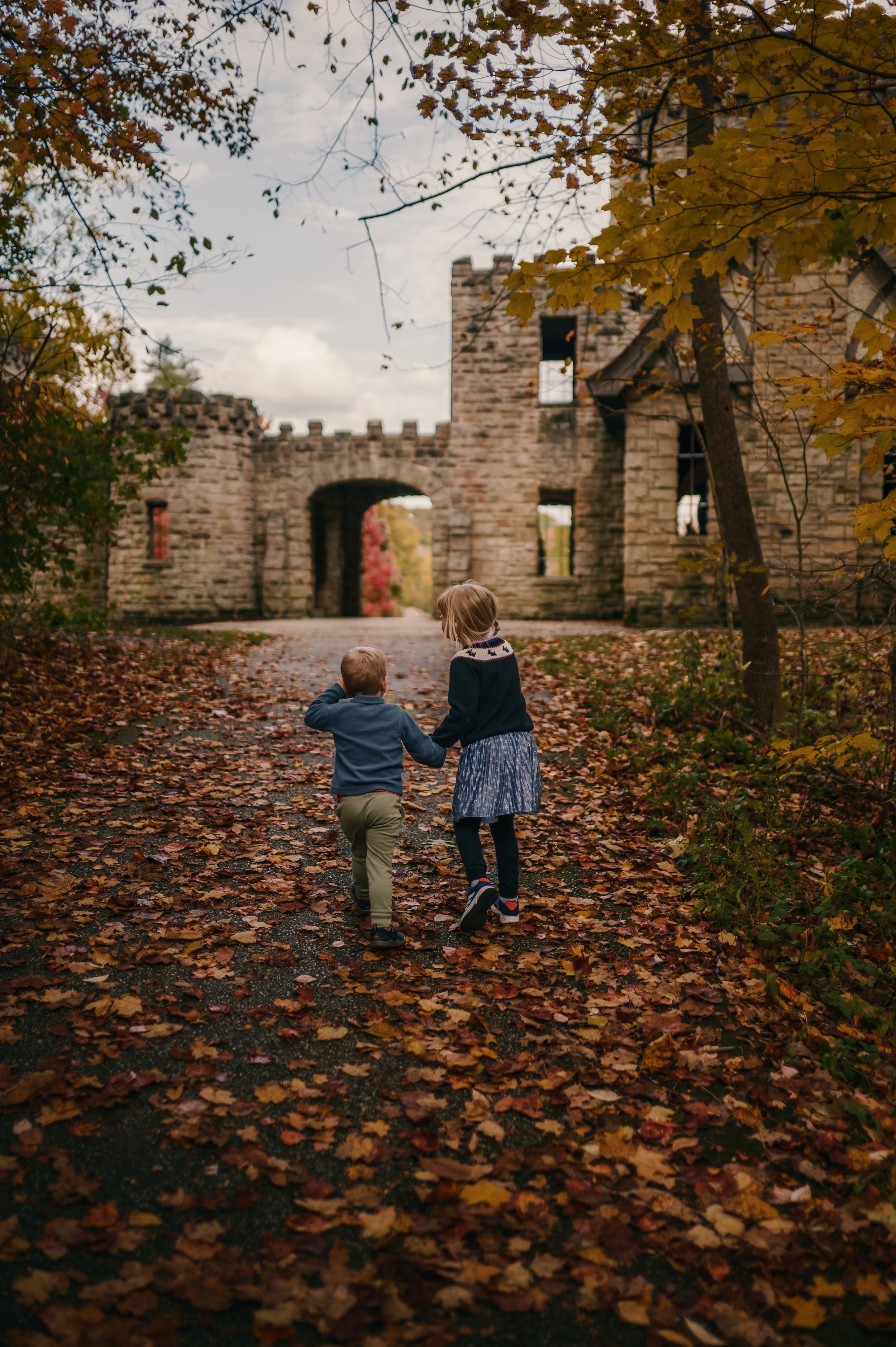 Two children holding hands walk toward an ancient stone castle ruins surrounded by colorful autumn foliage.