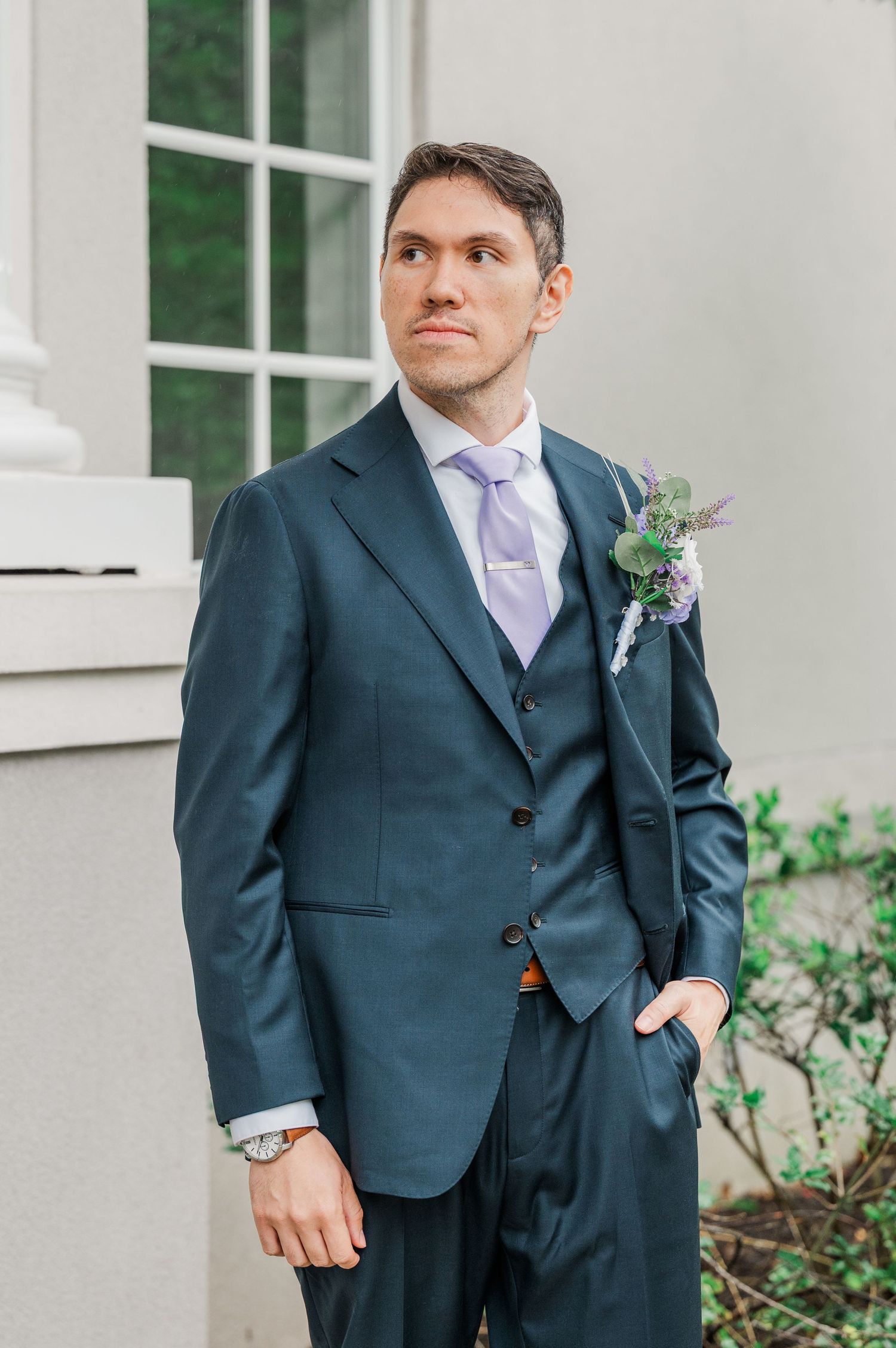 Groom in navy blue suit with lavender tie and floral boutonniere poses outdoors at wedding venue.