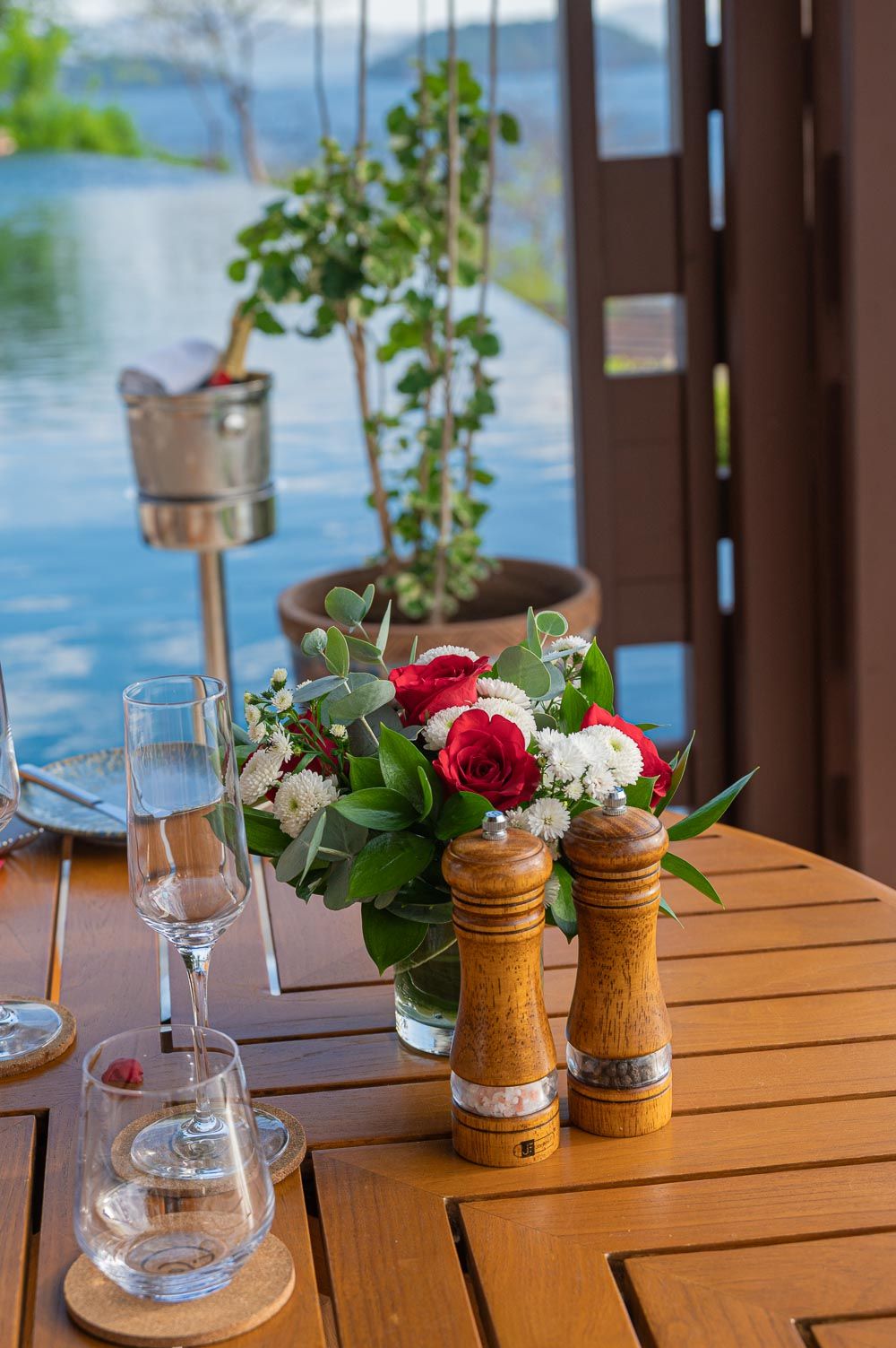 Elegant outdoor dining table with red and white roses, wooden pepper mills, wine glasses, and a champagne bucket near a pool.