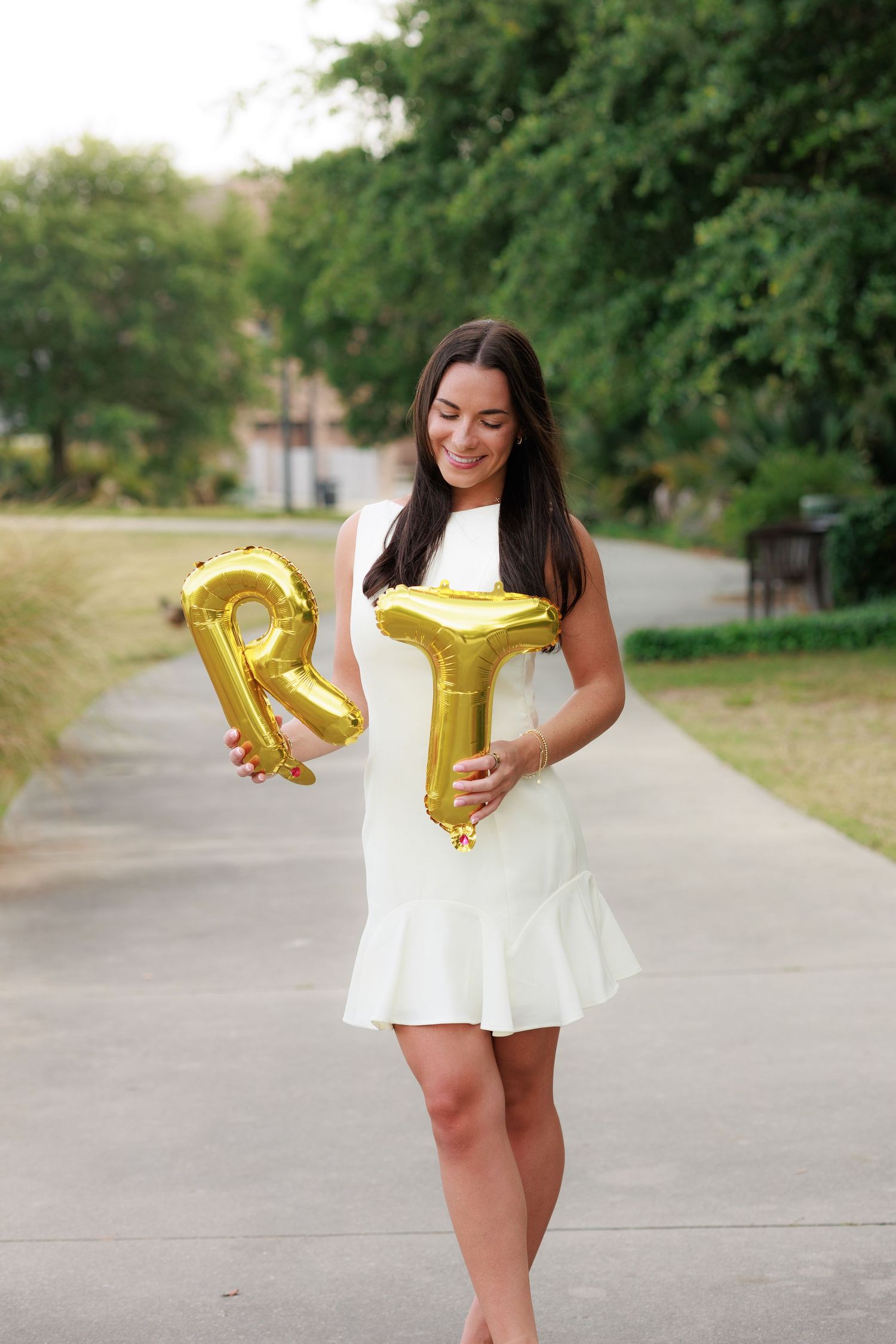 Young woman in white dress holding gold letter R and T balloons on a tree-lined pathway.