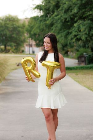 Woman in white dress holding gold letter R and T balloons on a tree-lined path outdoors.