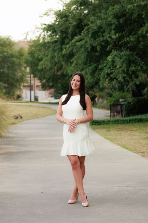 Young woman in white sleeveless mini dress and nude heels smiling outdoors on a pathway surrounded by green trees.