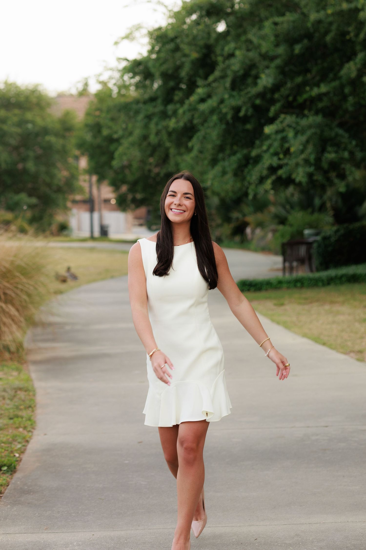 Smiling young woman in white sleeveless dress walking on a tree-lined pathway.