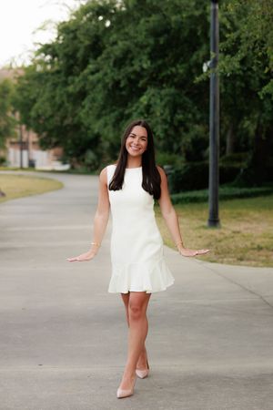 Young woman in white sleeveless dress and nude heels smiling outdoors on a path surrounded by green trees.