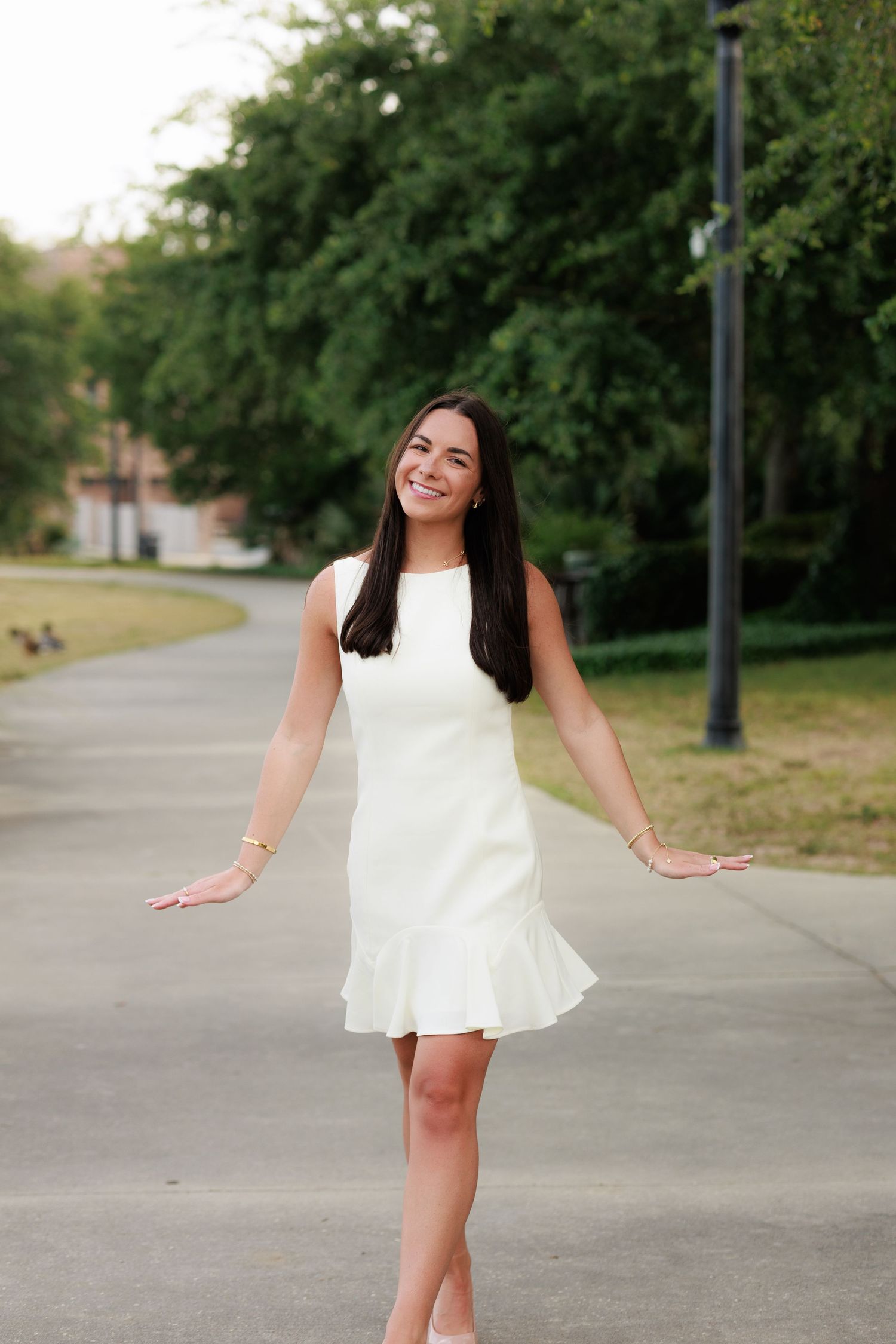 Young woman in white dress smiling with arms out on a tree-lined path.