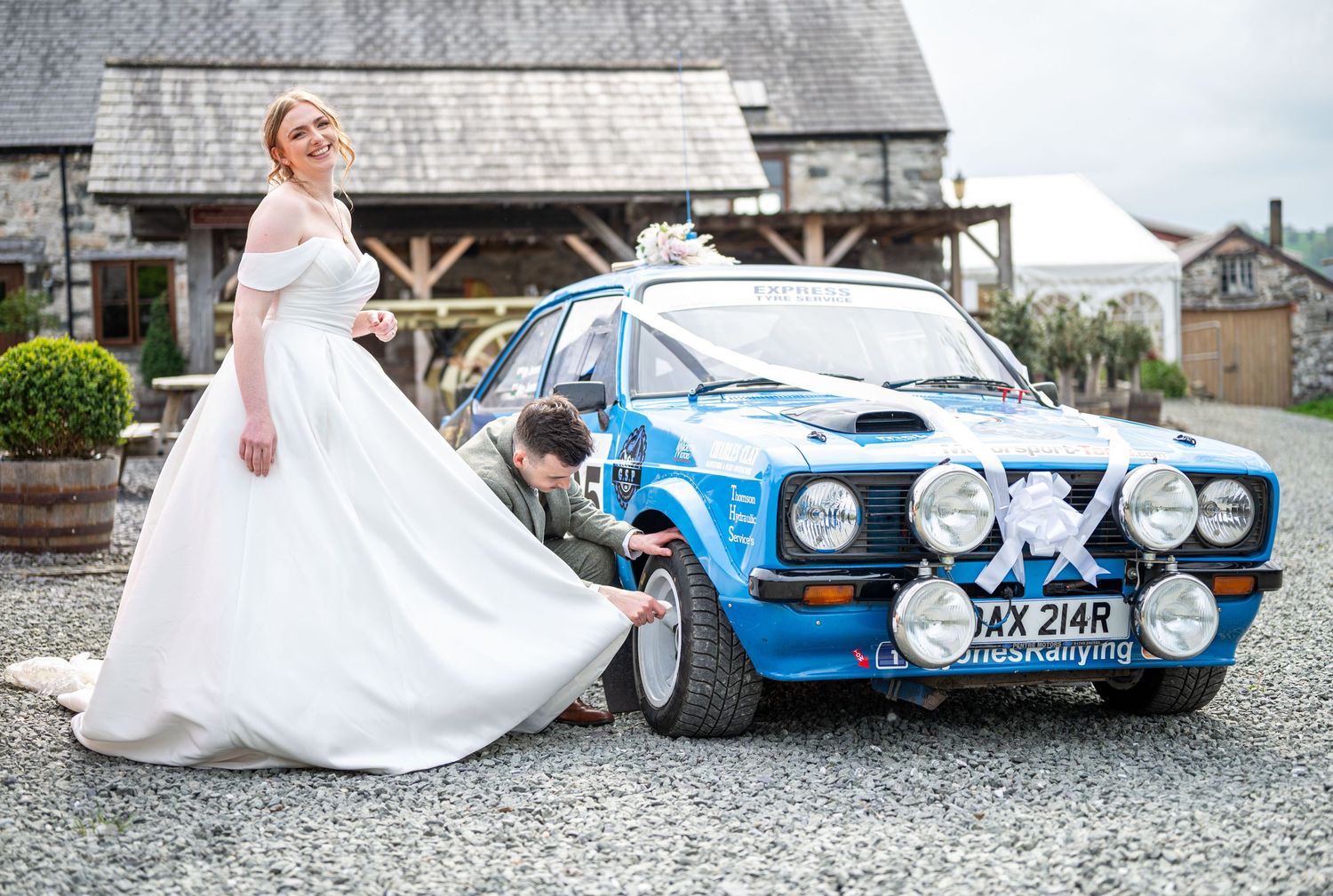 Bride in white gown poses beside a blue rally Ford Escort wedding car while groom adjusts decorations.