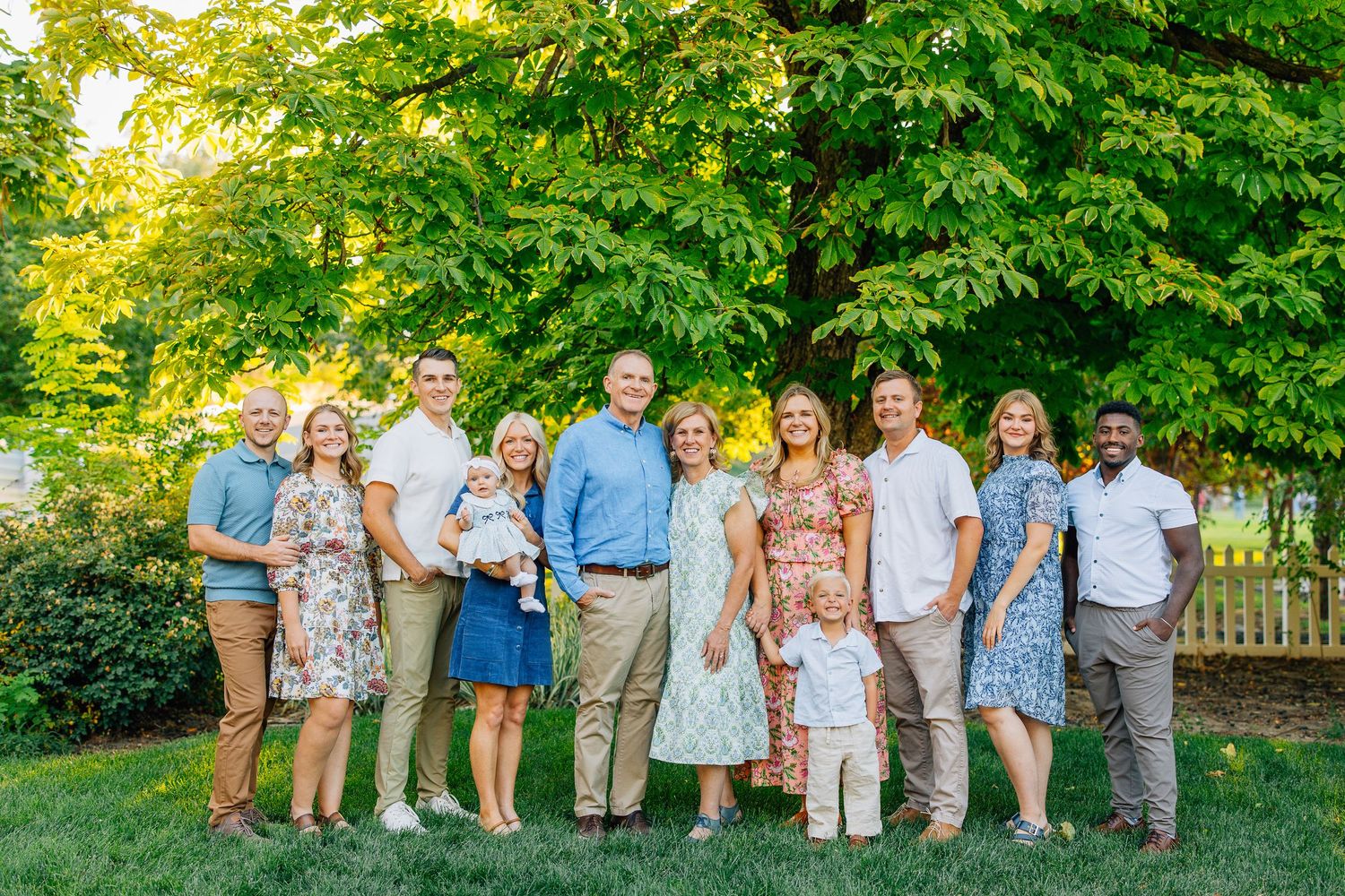 A large family group smiling outdoors on green grass with lush trees in the background on a sunny day. New York Family Photographer Saratoga