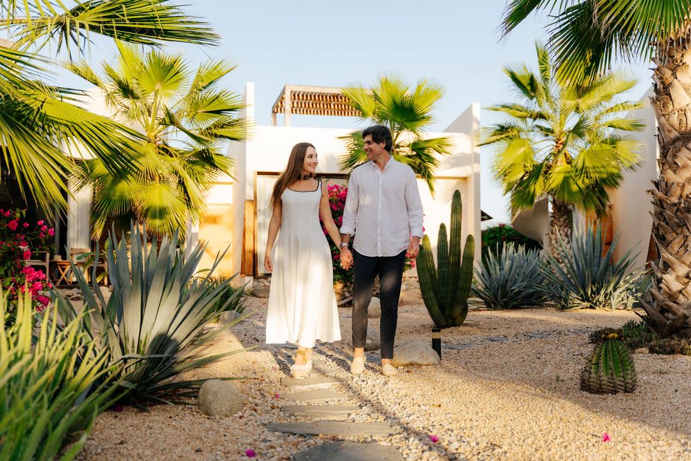 Couple walks hand-in-hand through a desert garden with palm trees and cacti near a white villa.