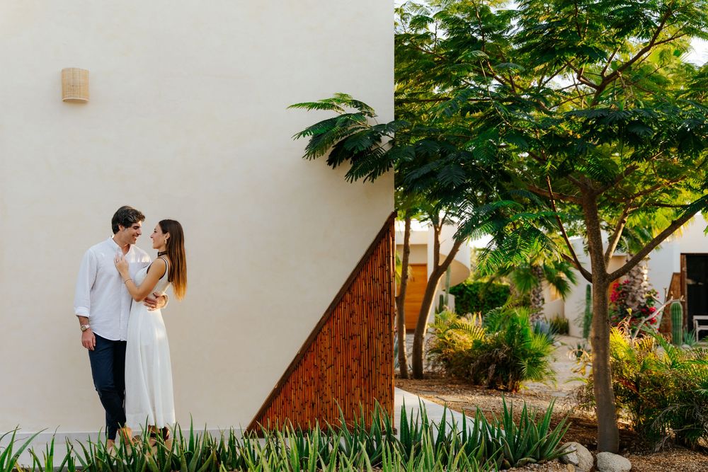 Couple embracing near modern white building surrounded by lush green trees and tropical plants in outdoor setting.