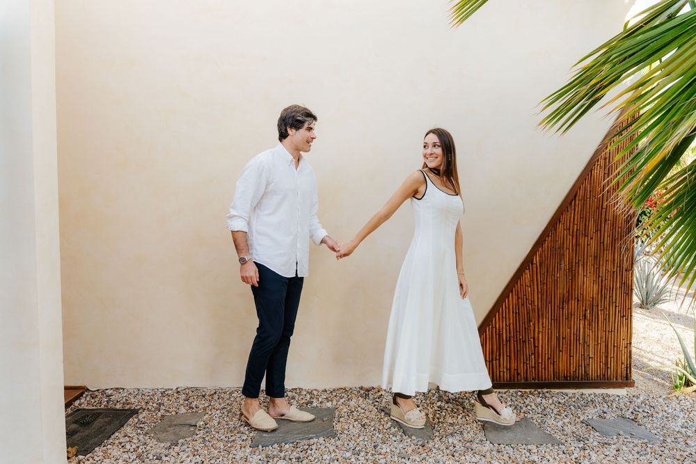 Couple holding hands in white outfits smiling outdoors near a palm tree and beige wall on a gravel path.
