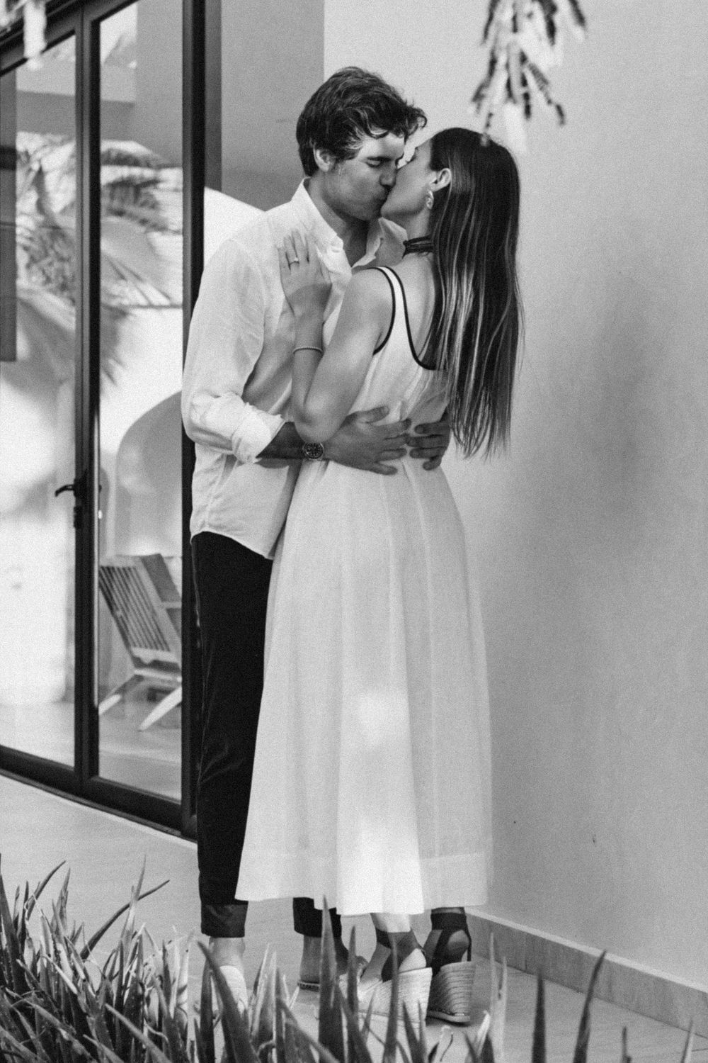 Black and white photo of a couple sharing a romantic kiss outdoors, woman in white dress, man in white shirt.