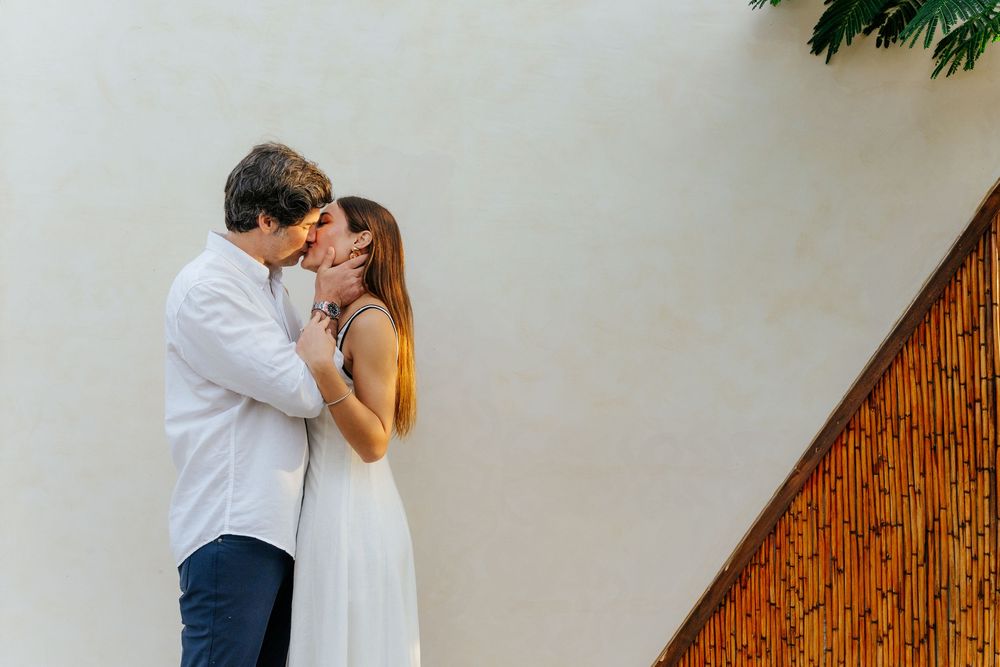 Romantic couple sharing a tender kiss outdoors against a white wall with tropical greenery.