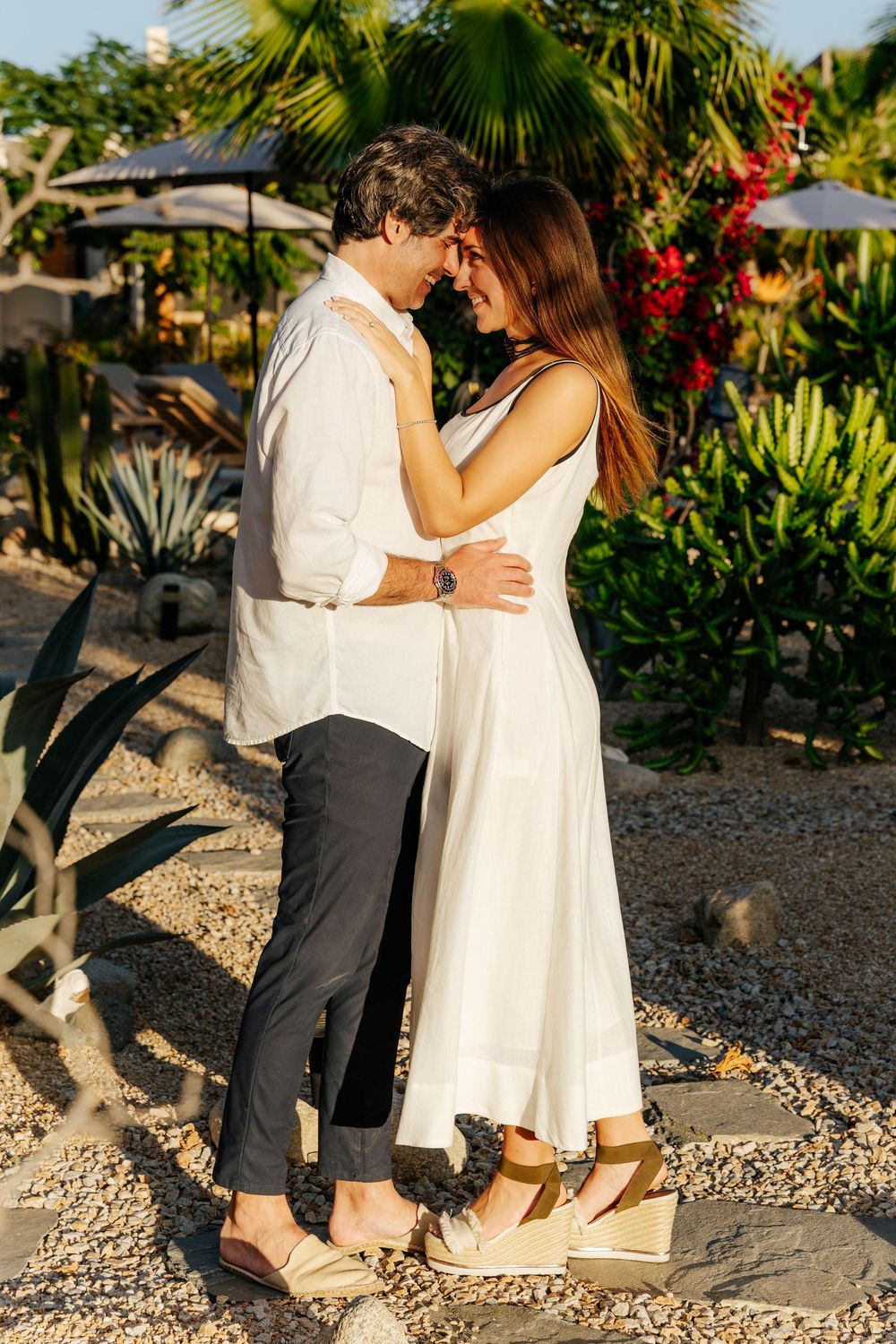 Romantic couple embracing in a tropical garden, woman in white dress, man in white shirt, surrounded by lush greenery.