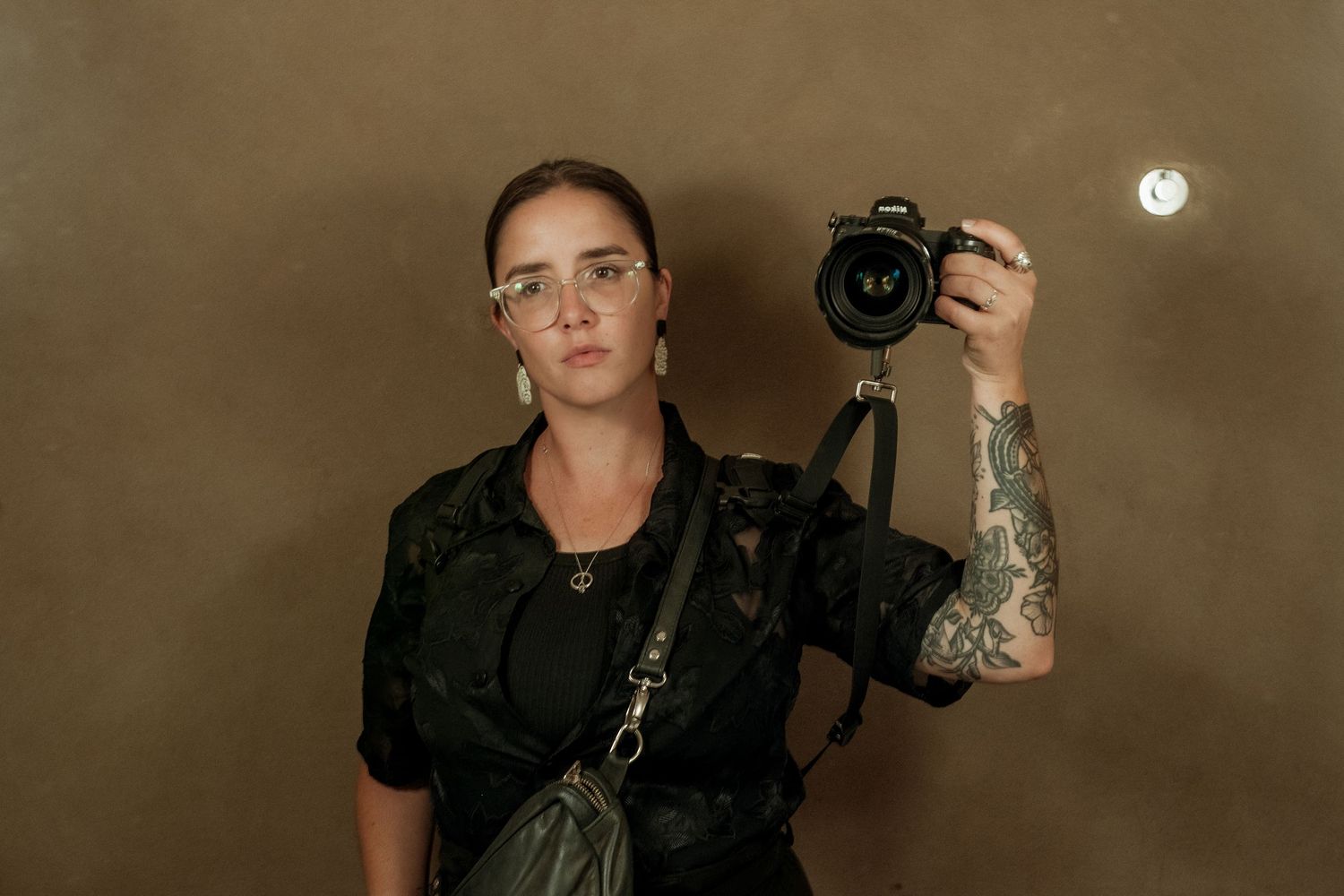 Tattooed female photographer holding a DSLR camera, wearing safety glasses and black outfit against brown wall.