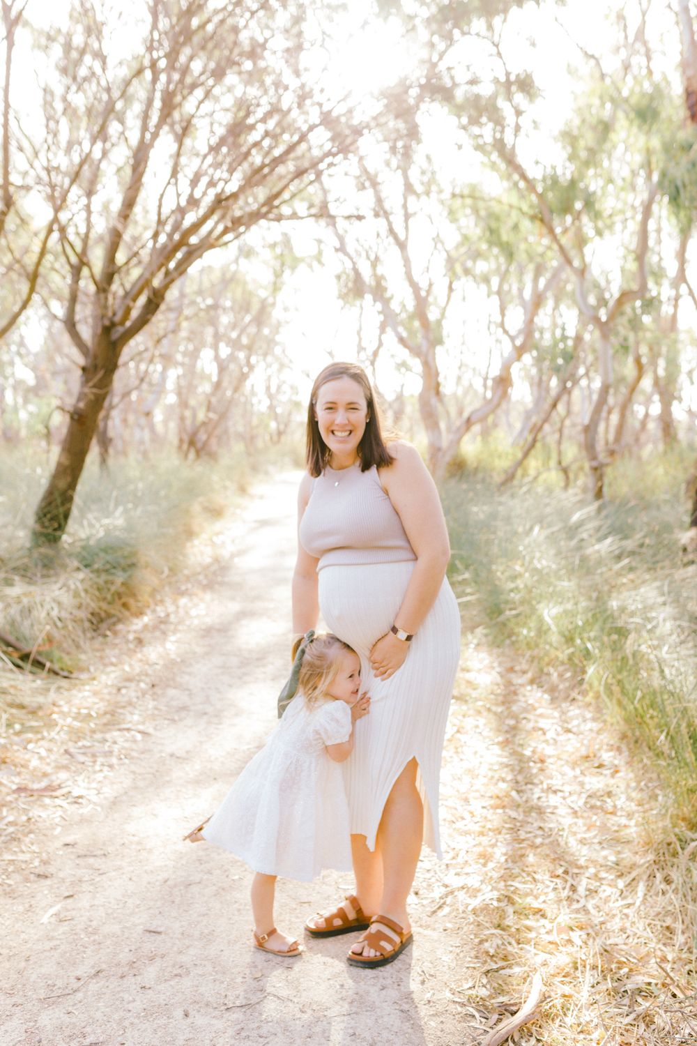 Pregnant woman in white skirt smiles outdoors as toddler girl hugs her baby bump on a sunlit forest path.