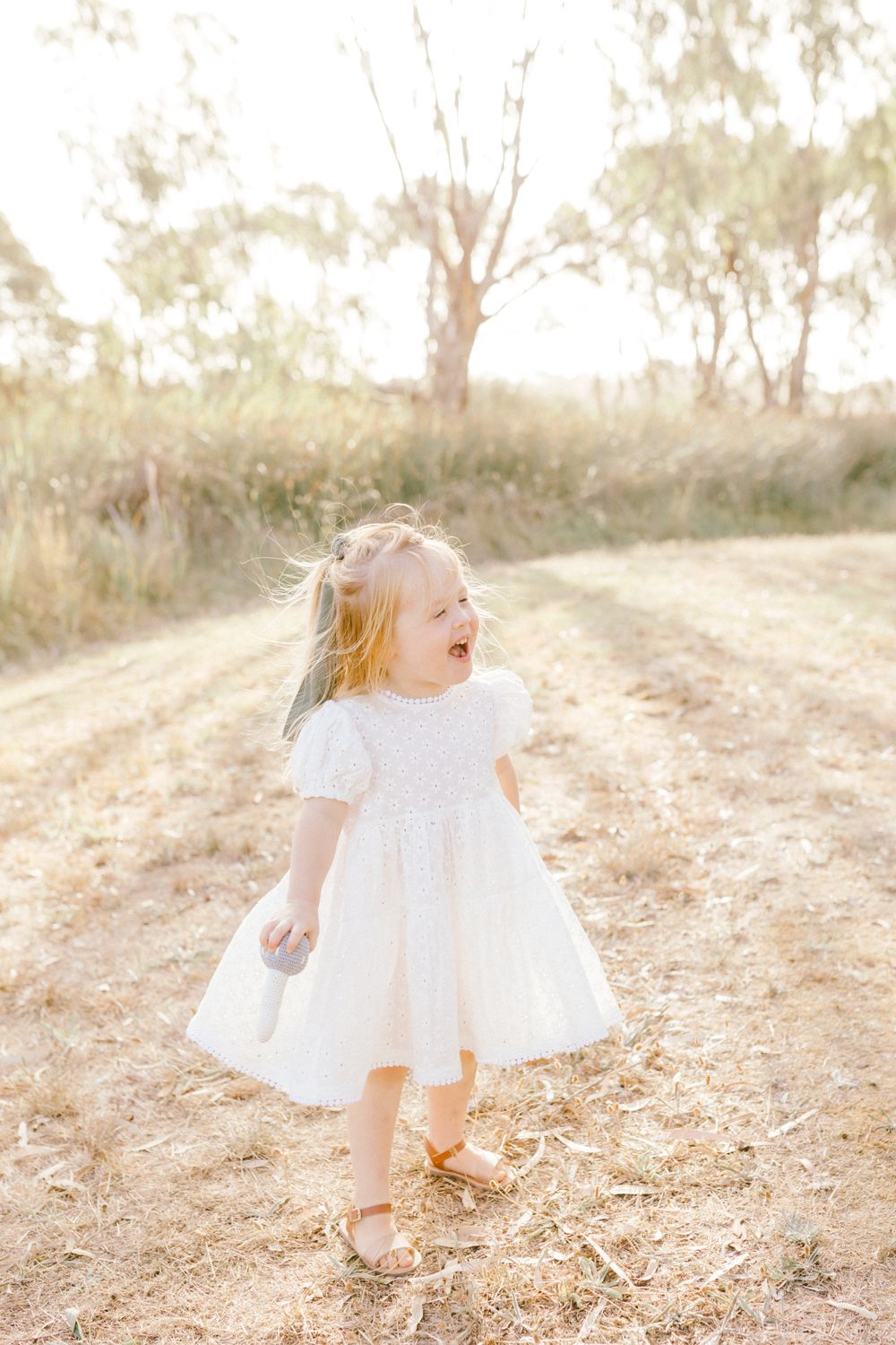 A joyful toddler girl in a white dress and gold sandals laughs outdoors on a sunny dirt path surrounded by dry grass.