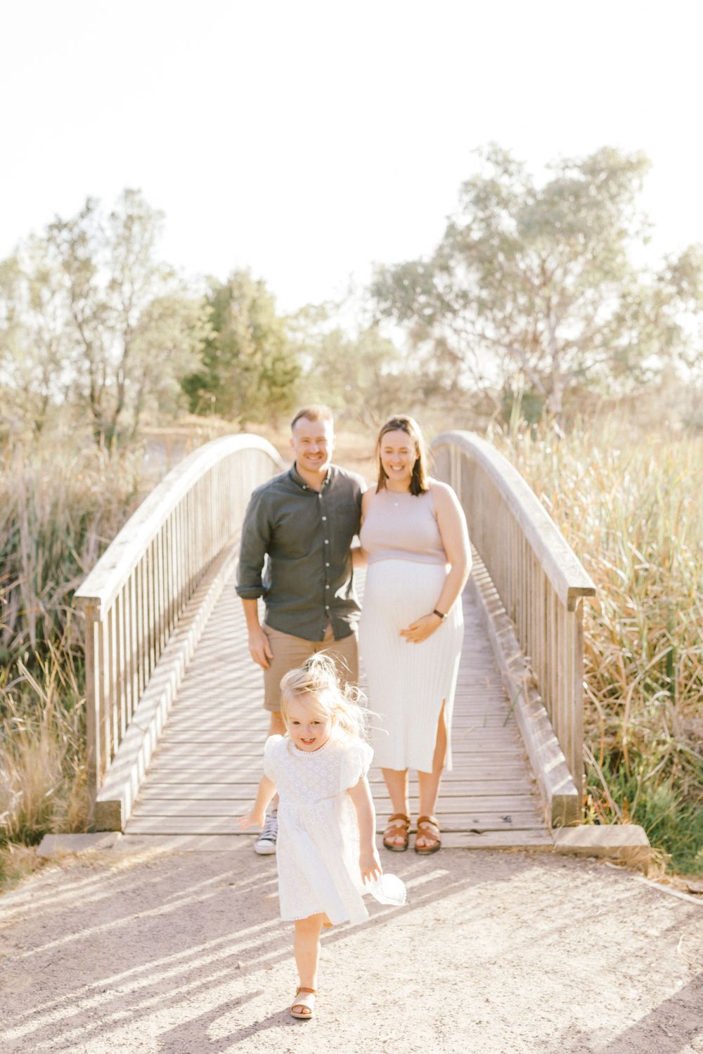 Pregnant woman and man stand on wooden bridge while young girl in white dress runs toward camera outdoors.