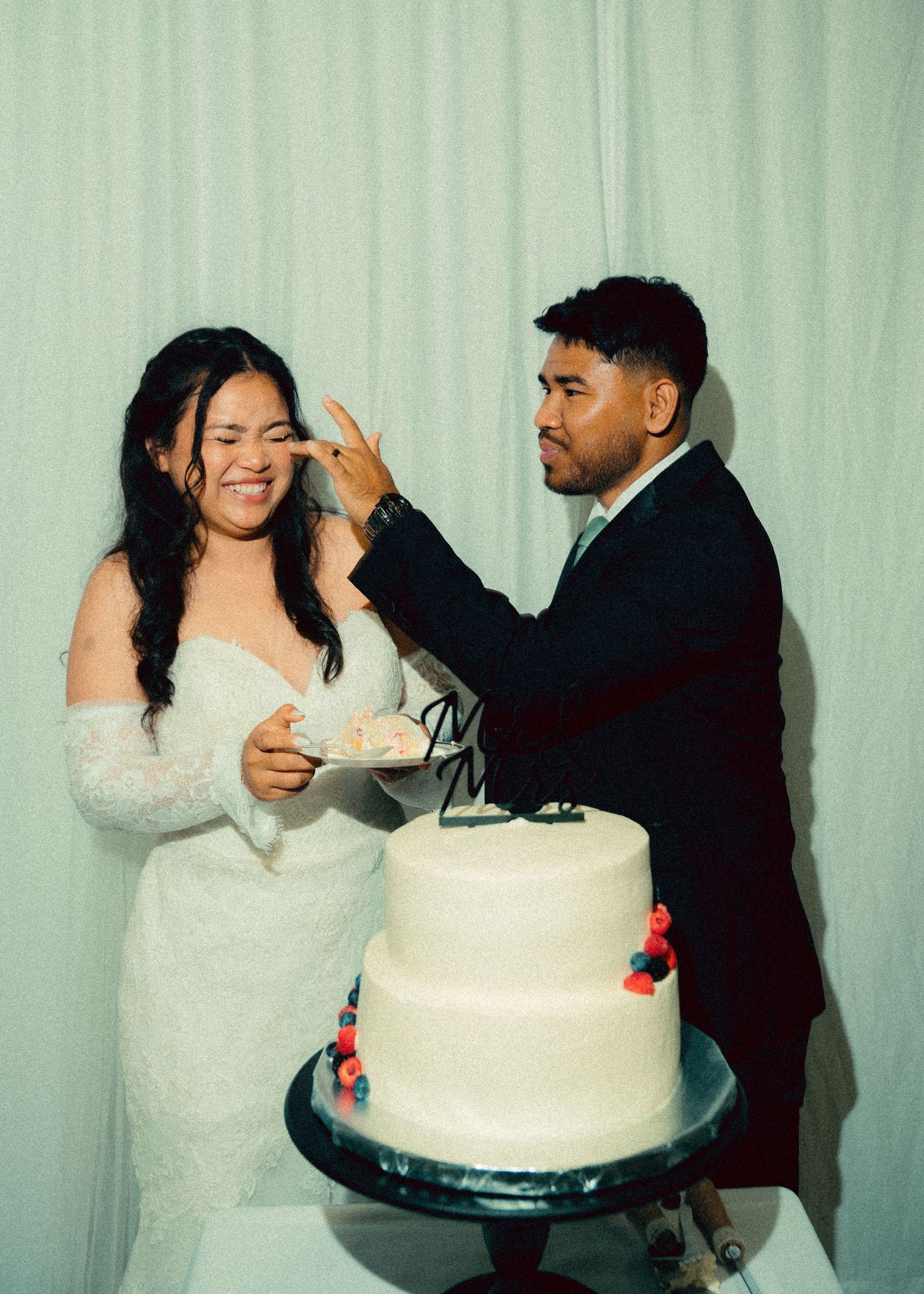 Laughing bride and groom smash wedding cake together in front of a white two-tiered decorated cake.