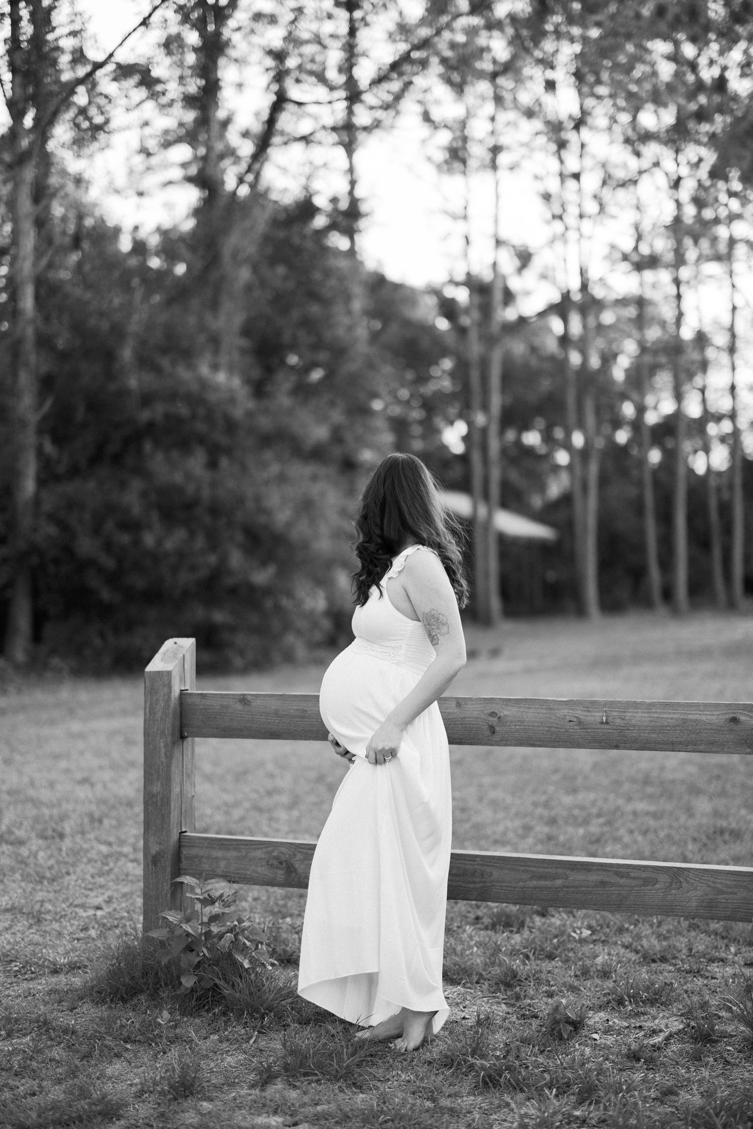 A pregnant mother in a flowing white dress stands on a sandy beach at sunset with rocky shore and ocean waves in background.