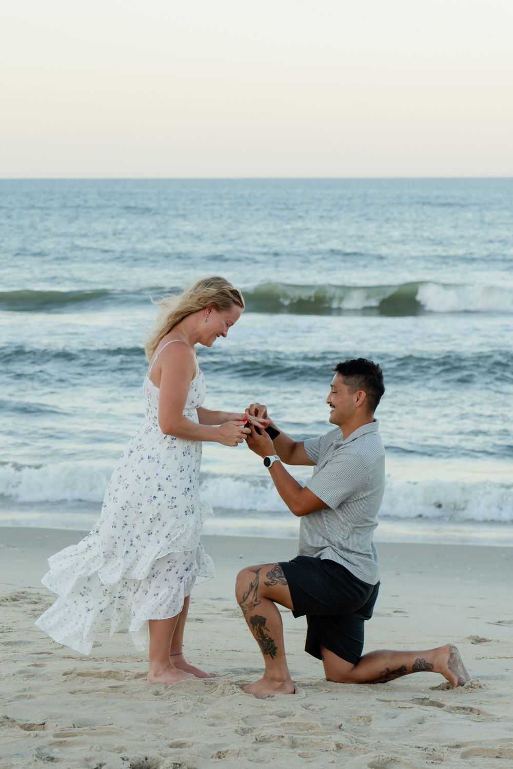 Man kneeling on beach proposing to woman in white dress, ocean waves in background at sunset.