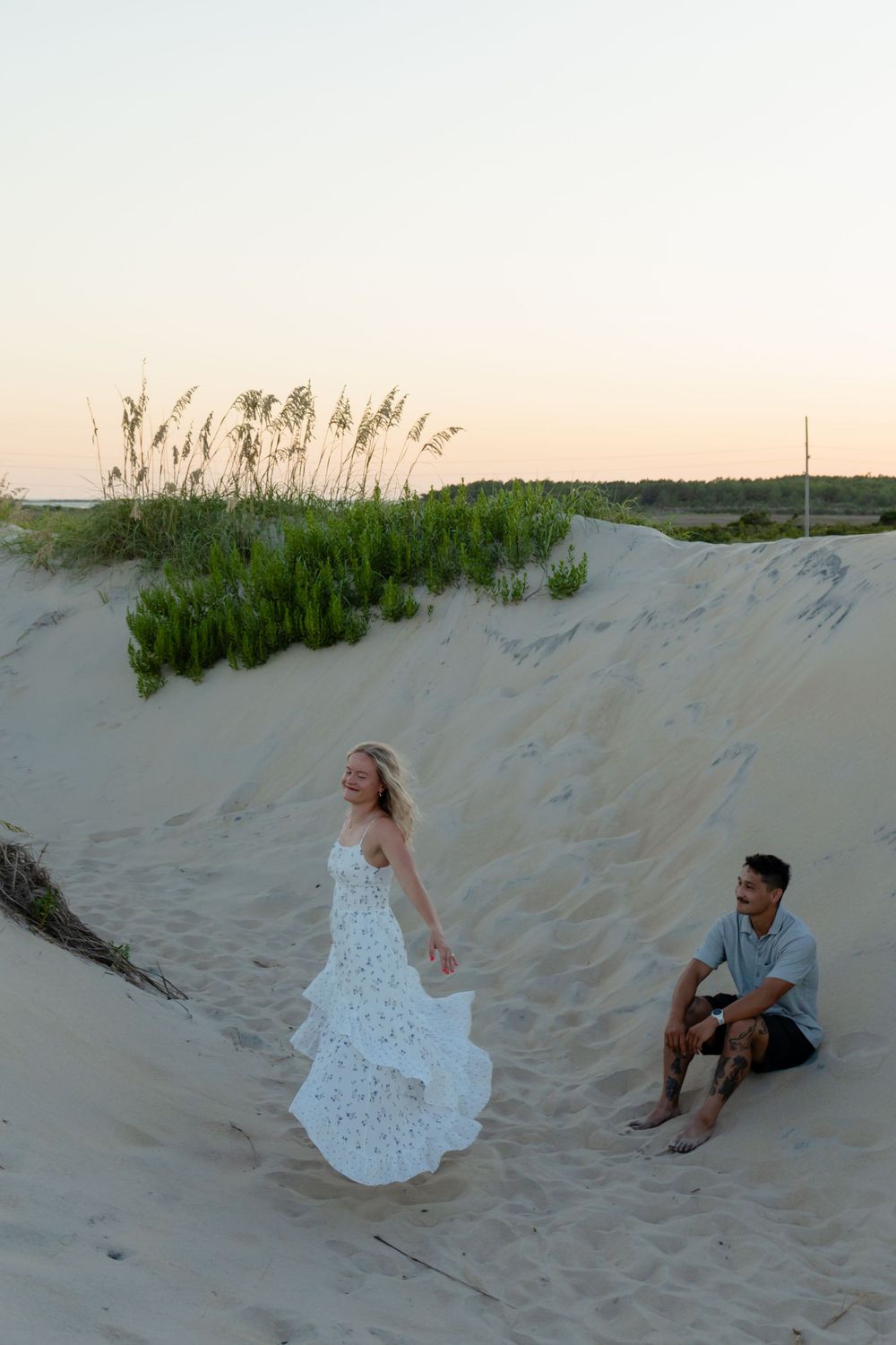 Couple on sandy dunes at sunset, woman in white lace dress twirling while man sits smiling beside beach grass.