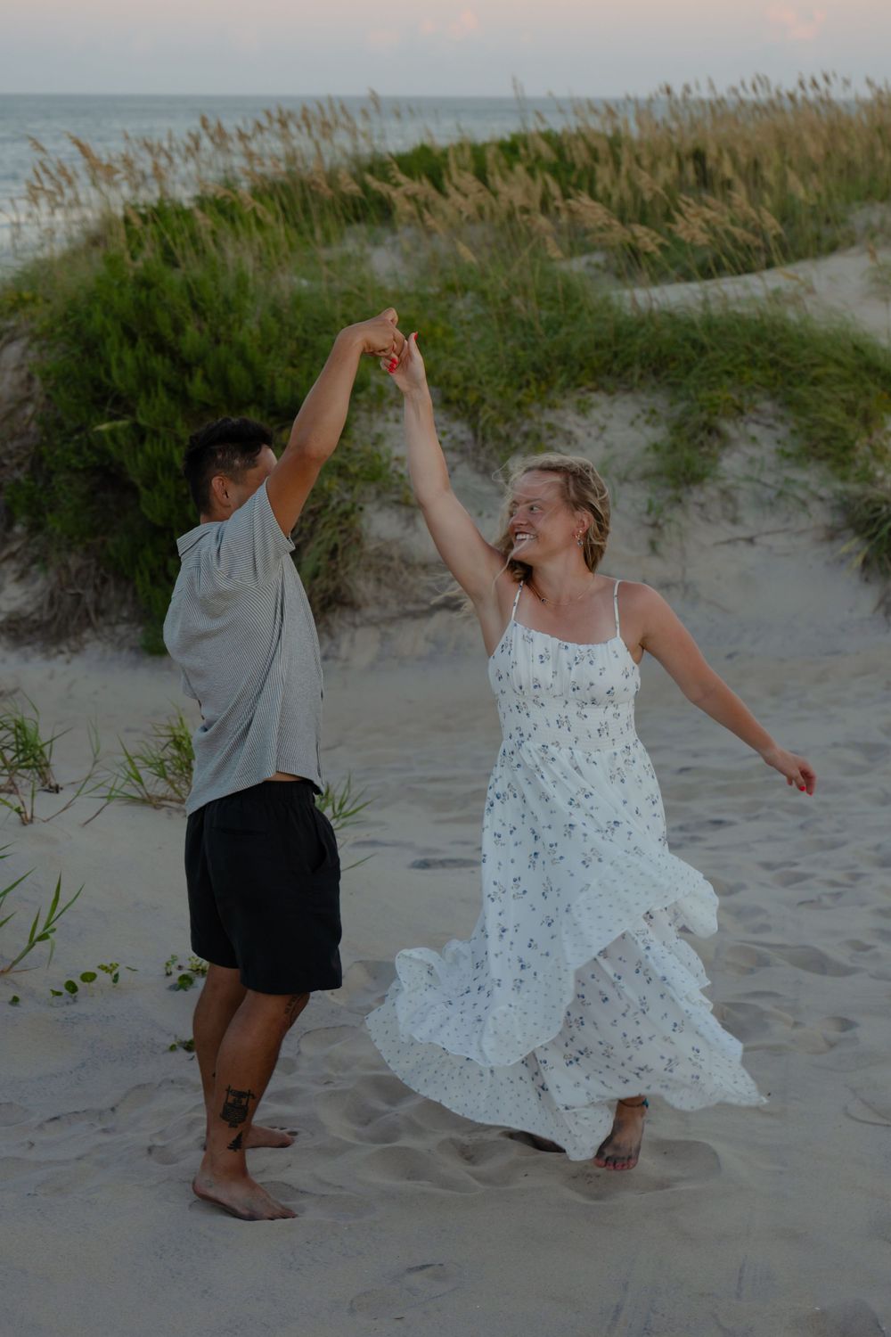 Couple dancing joyfully on sandy beach at dusk, woman in white floral dress twirling, ocean and dunes in background.