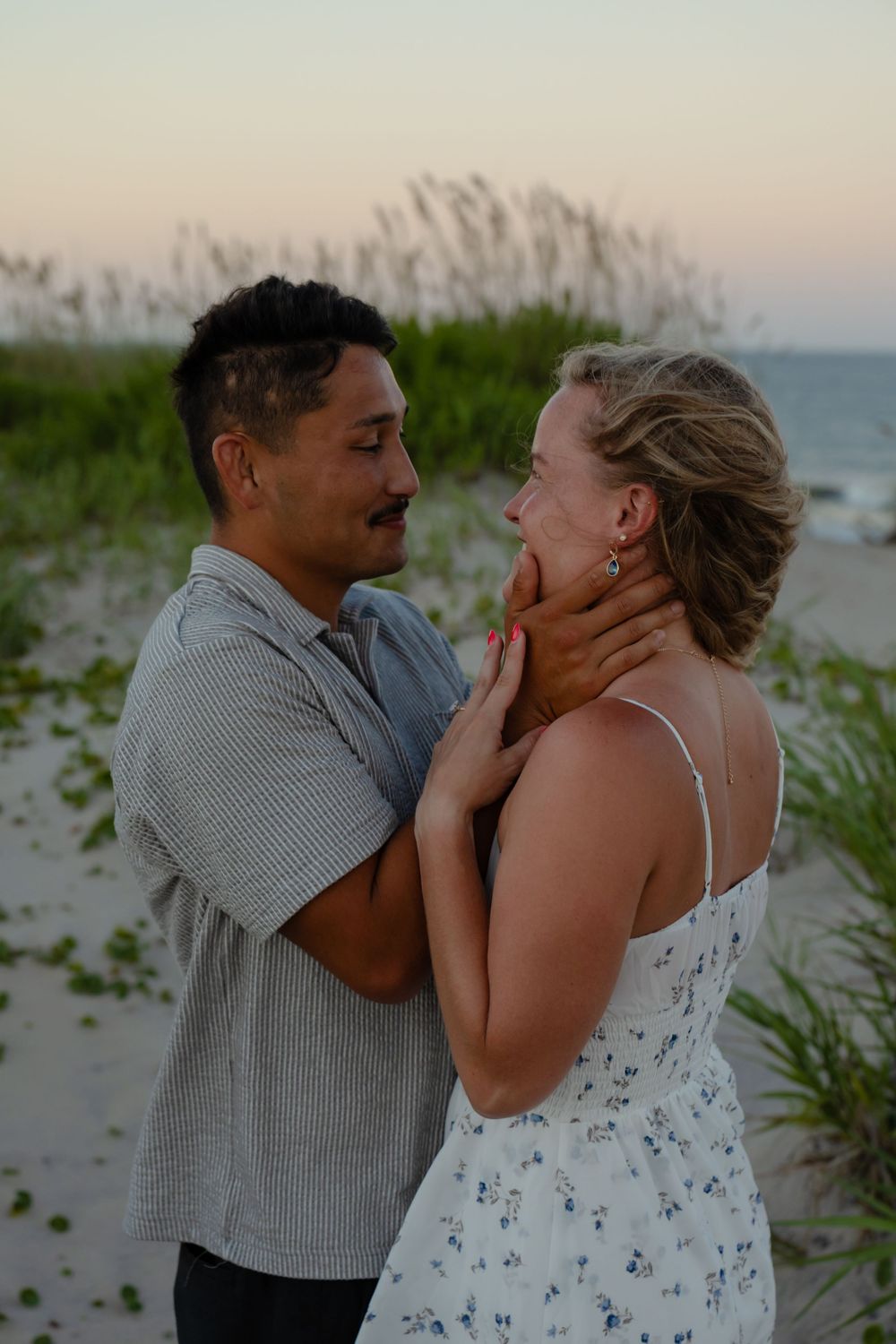 Couple sharing a romantic moment on a sandy beach at dusk, man gently holding woman's face near ocean dunes.