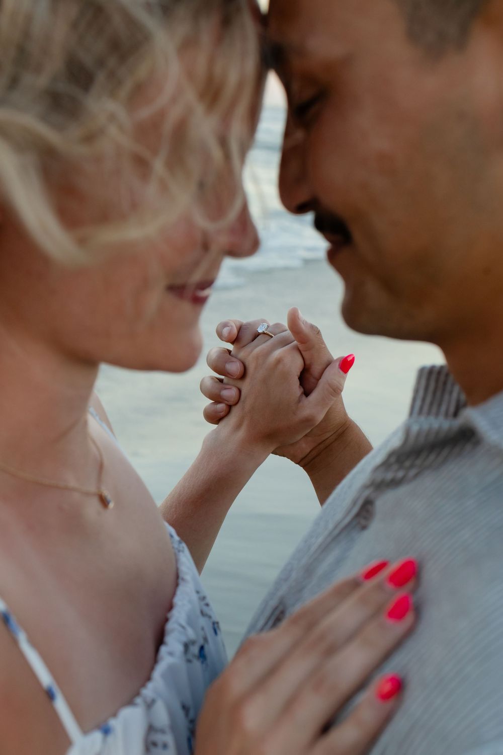 A romantic couple holds hands closely, the woman wearing a diamond ring and red nail polish, sharing an intimate moment together.