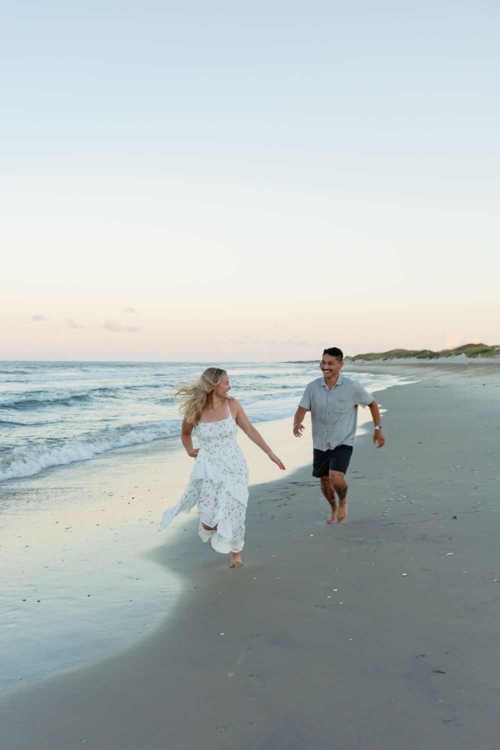 A couple holds hands and runs joyfully along a sandy beach at sunset, waves gently crashing behind them.