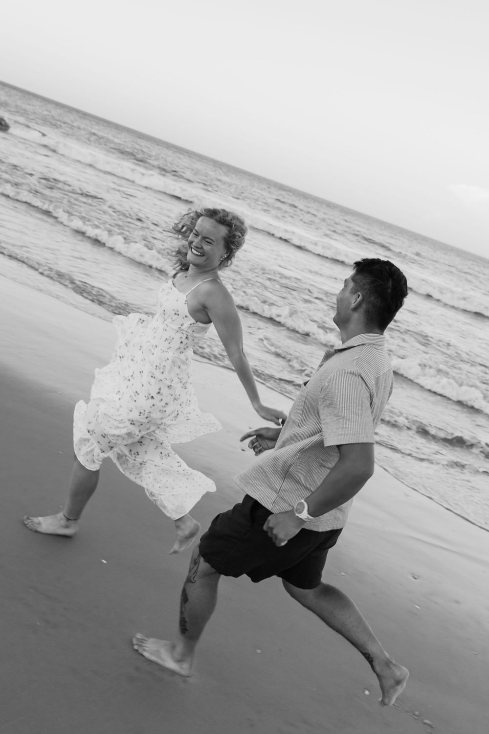 Black and white photo of a joyful couple running barefoot on a beach, holding hands, waves in the background.