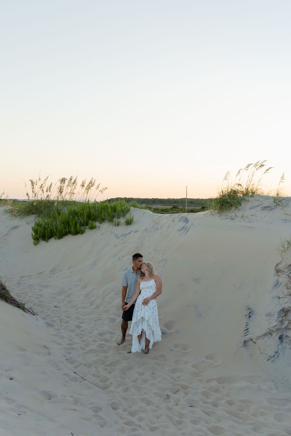Couple embracing on sandy beach dunes at sunset, woman in white dress, sea oats in background.