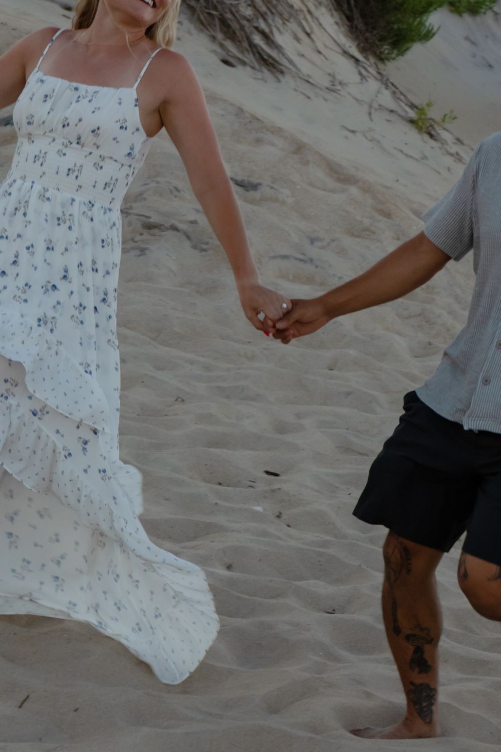 Couple holding hands on sandy beach, woman in floral maxi dress, man in gray shirt and black shorts with tattoo.