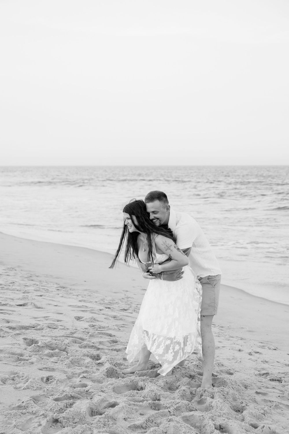 Black and white photo of a couple embracing and laughing on a sandy beach with ocean waves in the background.
