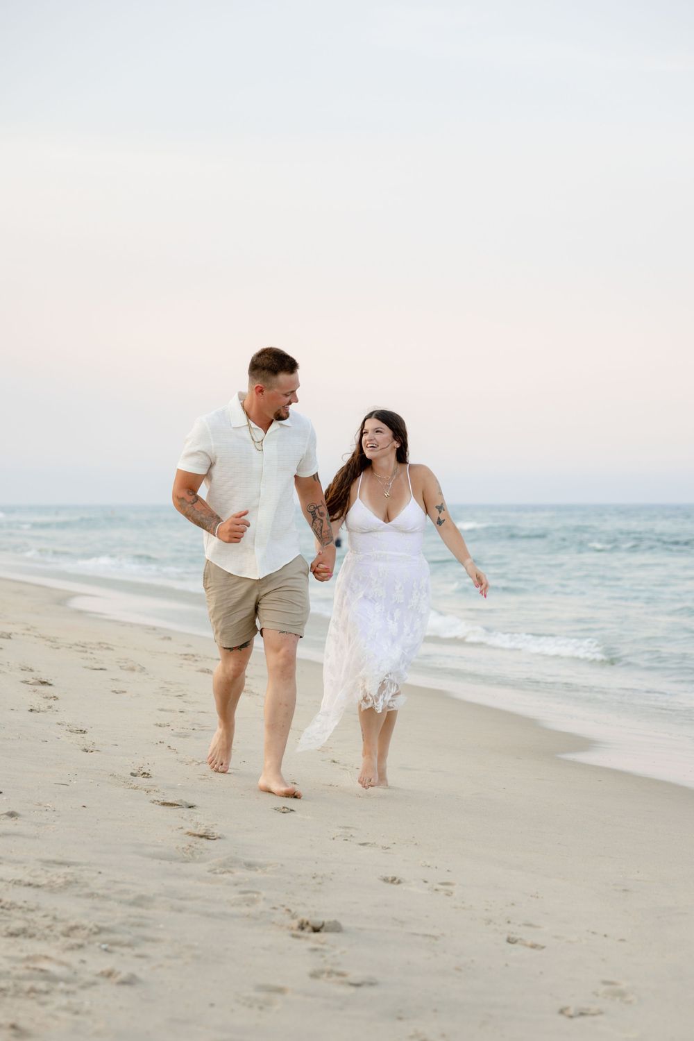 A couple in white outfits walks hand-in-hand along a sandy beach at sunset, smiling at each other.