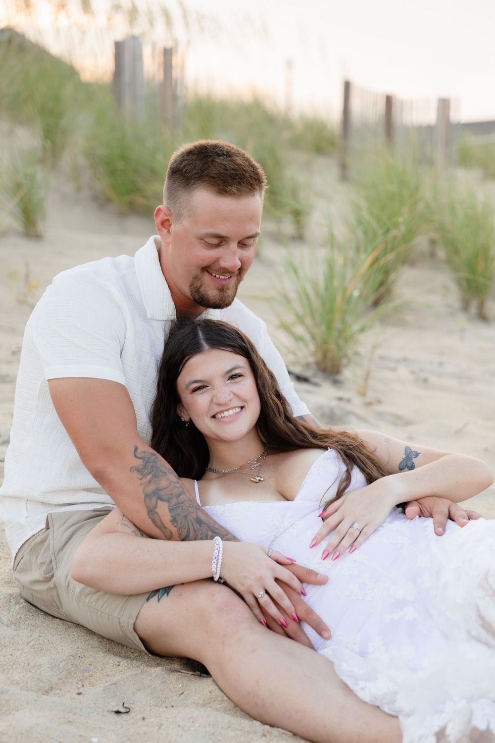 Couple sitting on sandy beach, man with tattoos embracing pregnant woman in white dress, smiling at baby bump.