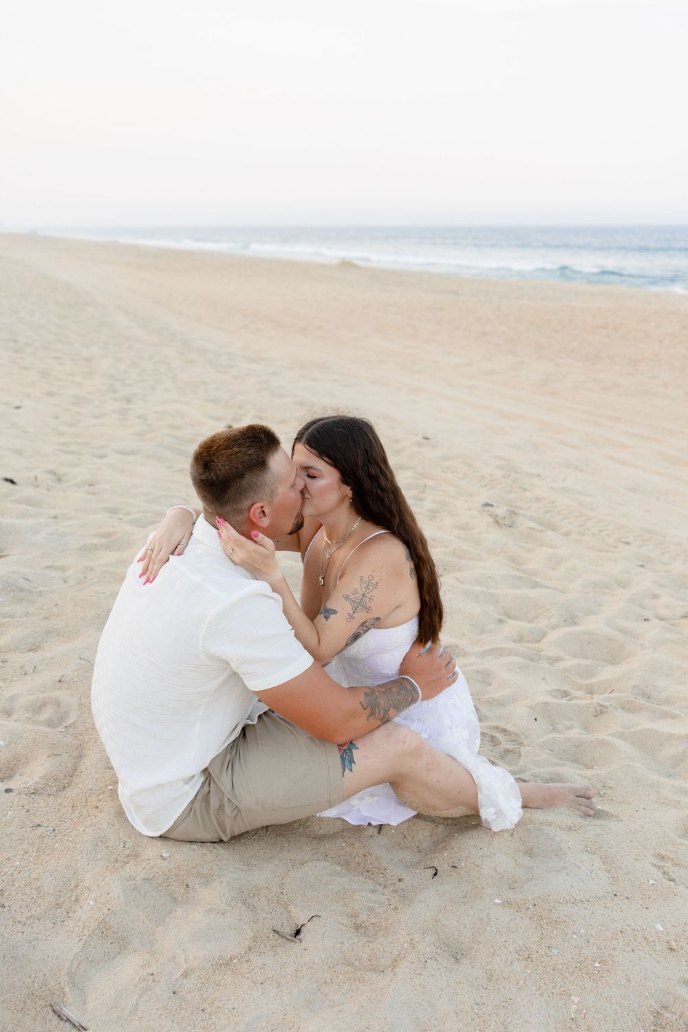 A couple shares a romantic kiss while sitting on a sandy beach, dressed in white outfits with ocean waves in the background.