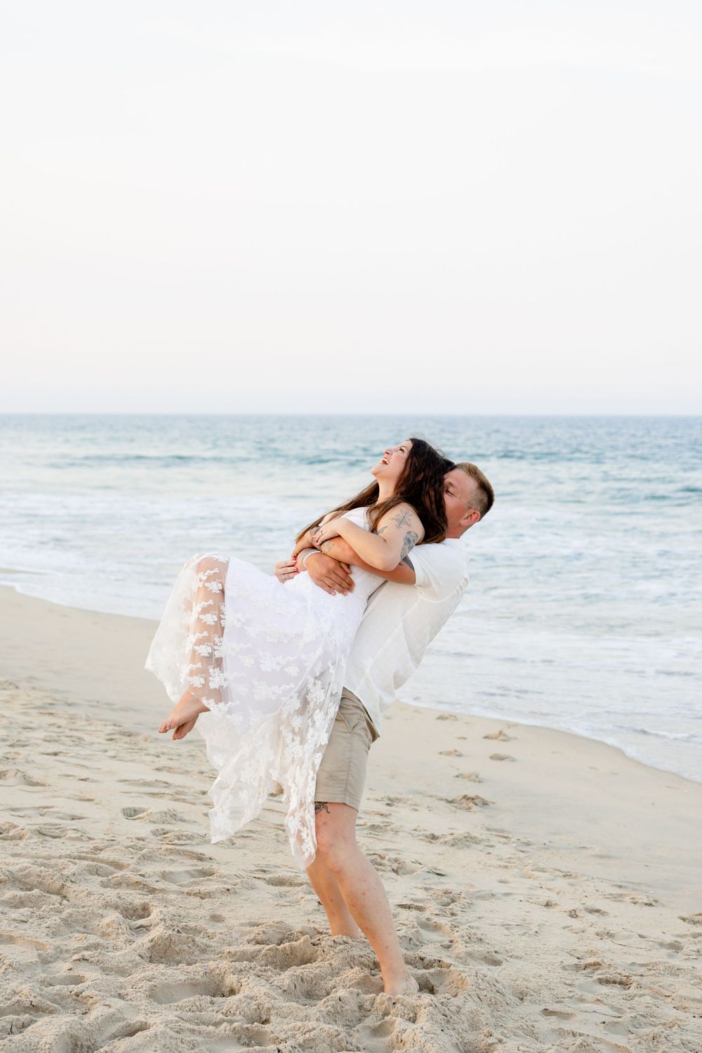 A joyful groom lifts his bride in a white lace dress on a sandy beach with ocean waves in the background.
