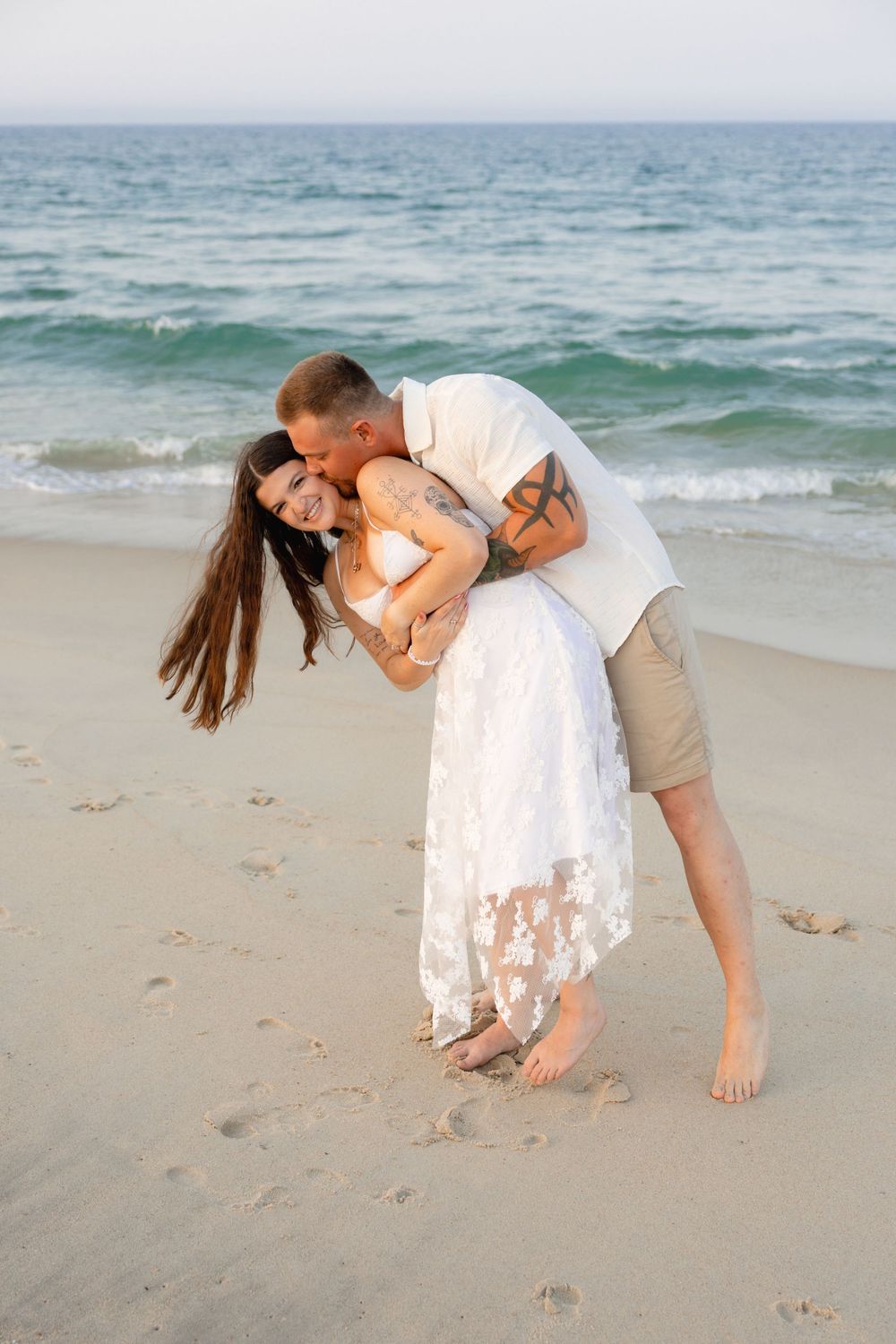 Couple embracing playfully on a sandy beach, woman in white lace dress laughing as man lifts her by the ocean.