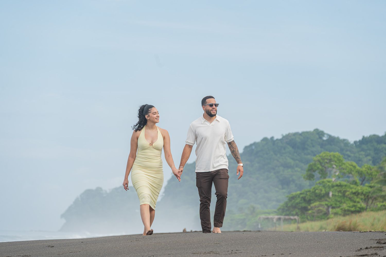 Couple holding hands walking on a black sand beach with lush green trees and misty ocean in the background.