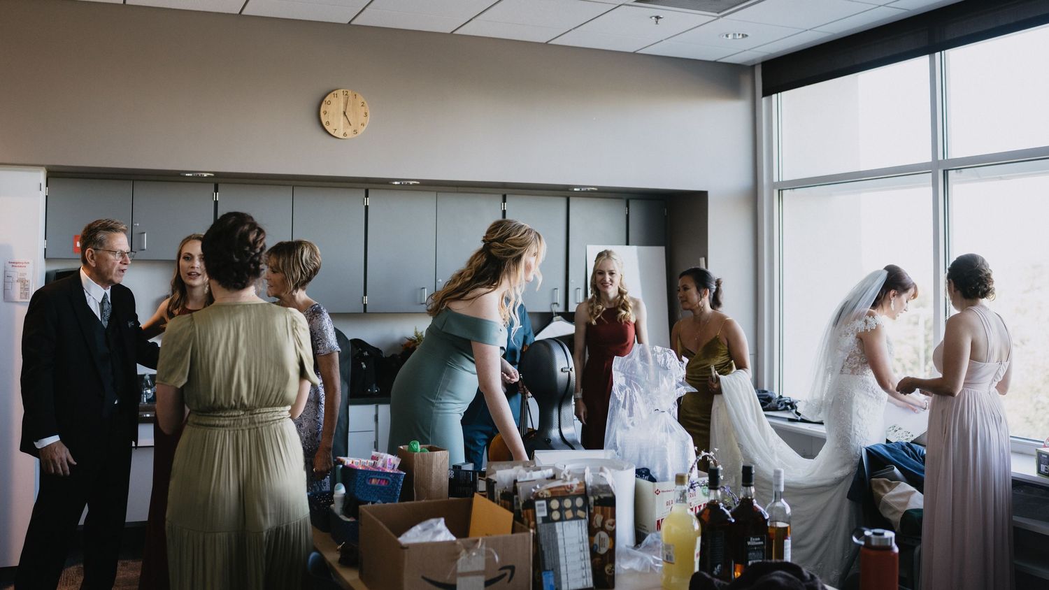 Wedding party preparing in a bright room, bridesmaids in sage green dresses surround the bride in her white gown.