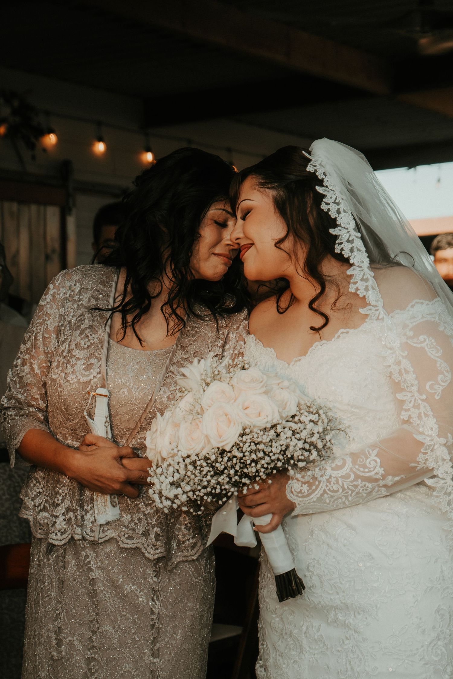 Bride in white lace gown and veil shares tender moment with guest in beige lace dress, holding white rose bouquet.