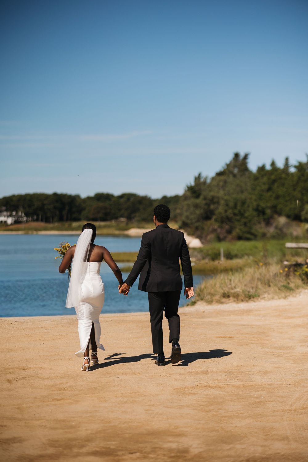 A bride and groom walk hand in hand along a sandy beach path near a calm lake on a sunny wedding day.