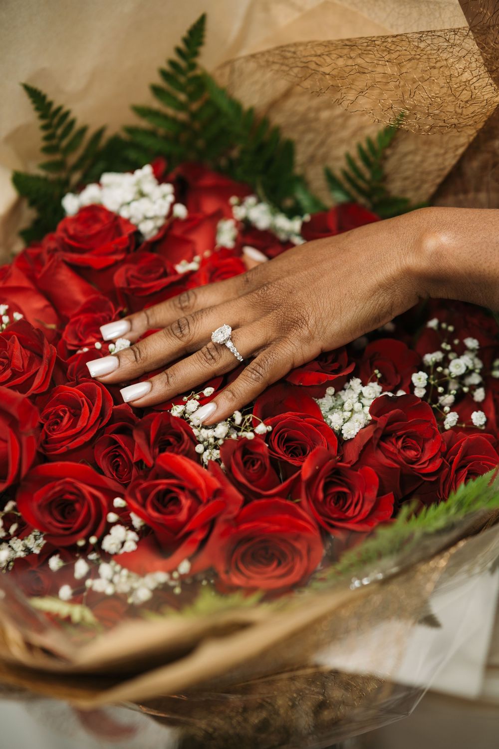 A hand with a diamond engagement ring resting on a bouquet of red roses with baby's breath and fern leaves.