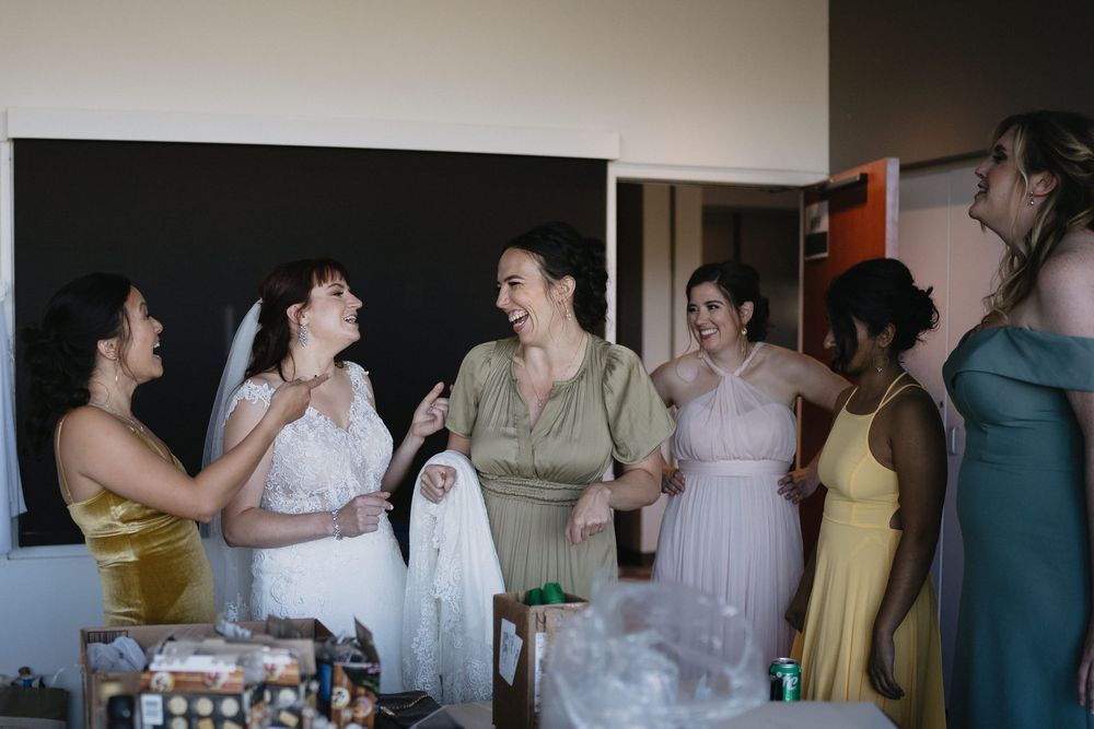 Bride laughing with bridesmaids in colorful dresses during wedding preparations indoors.